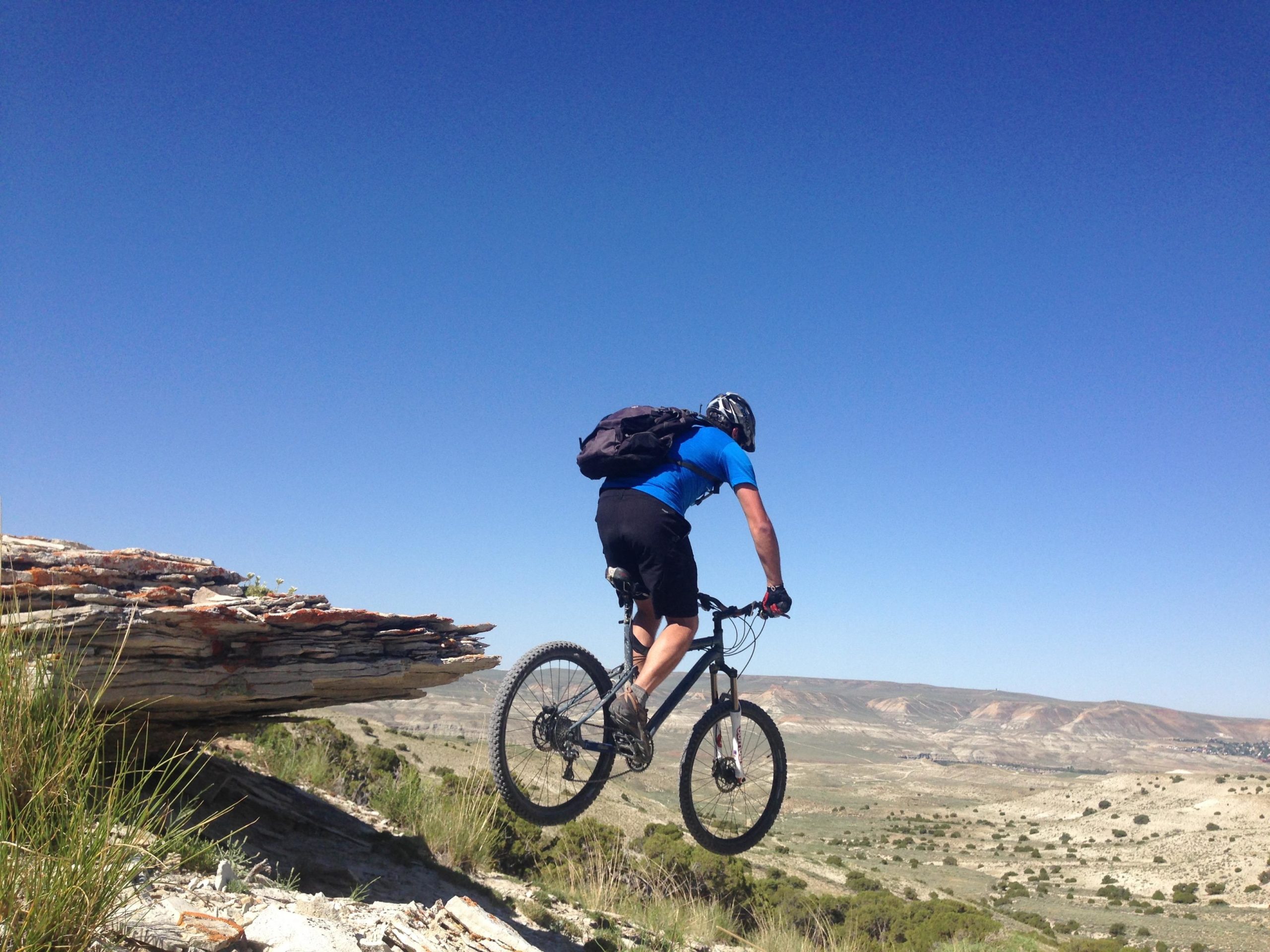 A mountain biker in a blue shirt and black shorts is jumping off a rocky ledge, with a panoramic view of a dry, hilly landscape and clear blue sky in the background. The bike is airborne, capturing the sense of adventure and excitement in outdoor sports. Brent & Mikes' Trail mountain bike trail.