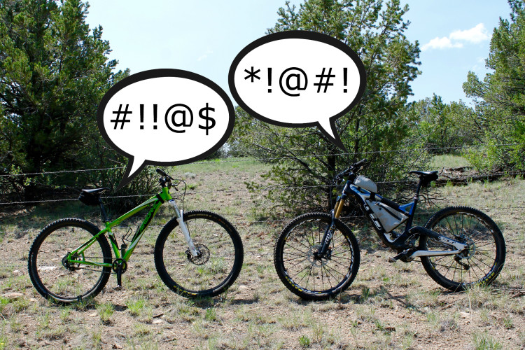 Two mountain bikes are positioned on a dirt surface, with bushes in the background. Each bike has a speech bubble above it containing symbols often used to represent swearing or strong reactions, suggesting a humorous or exaggerated conversation between the bikes.