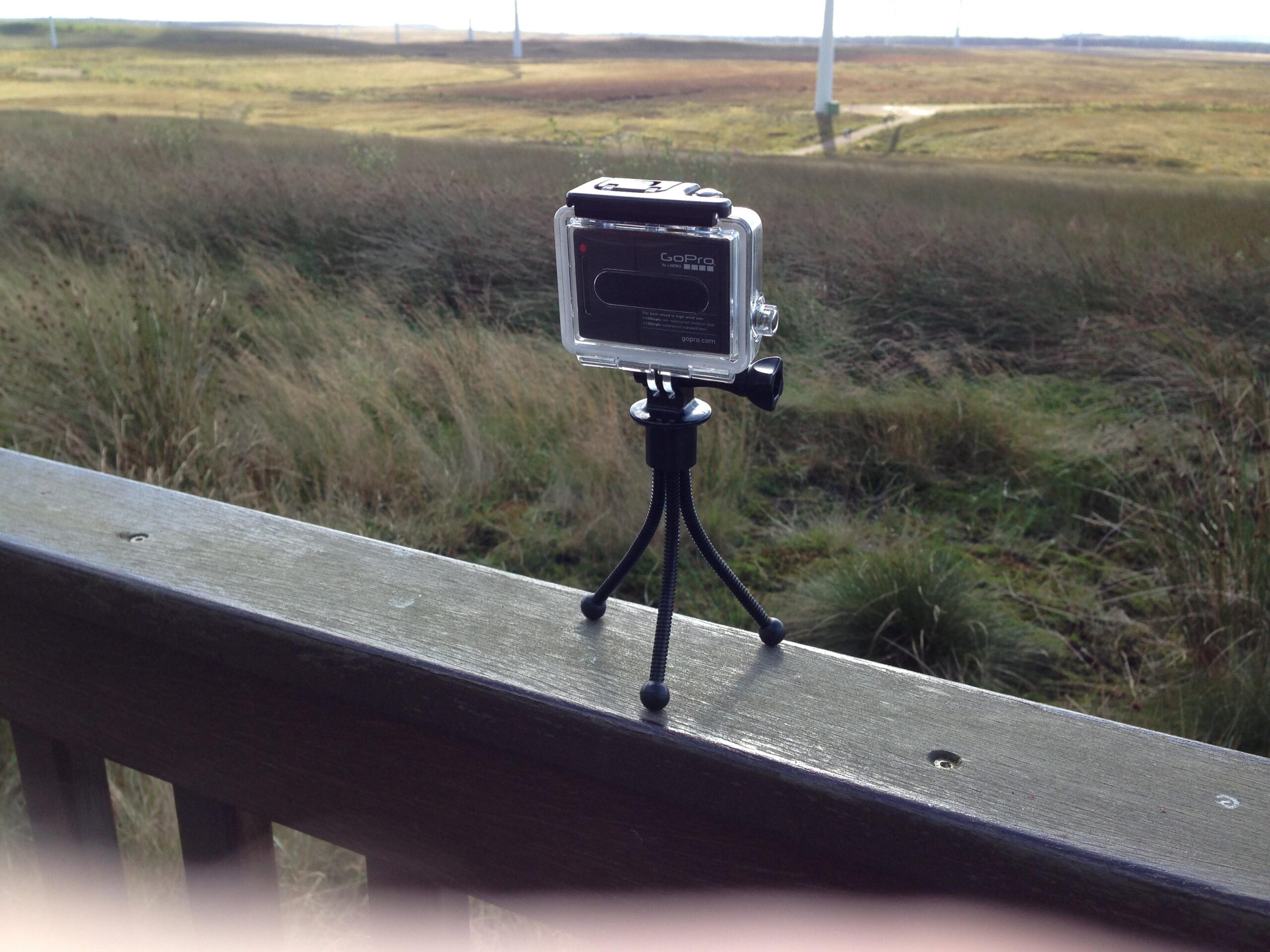 A GoPro camera mounted on a mini tripod is positioned on a wooden railing, overlooking a grassy landscape with wind turbines in the background. Whitelees mountain bike trail.