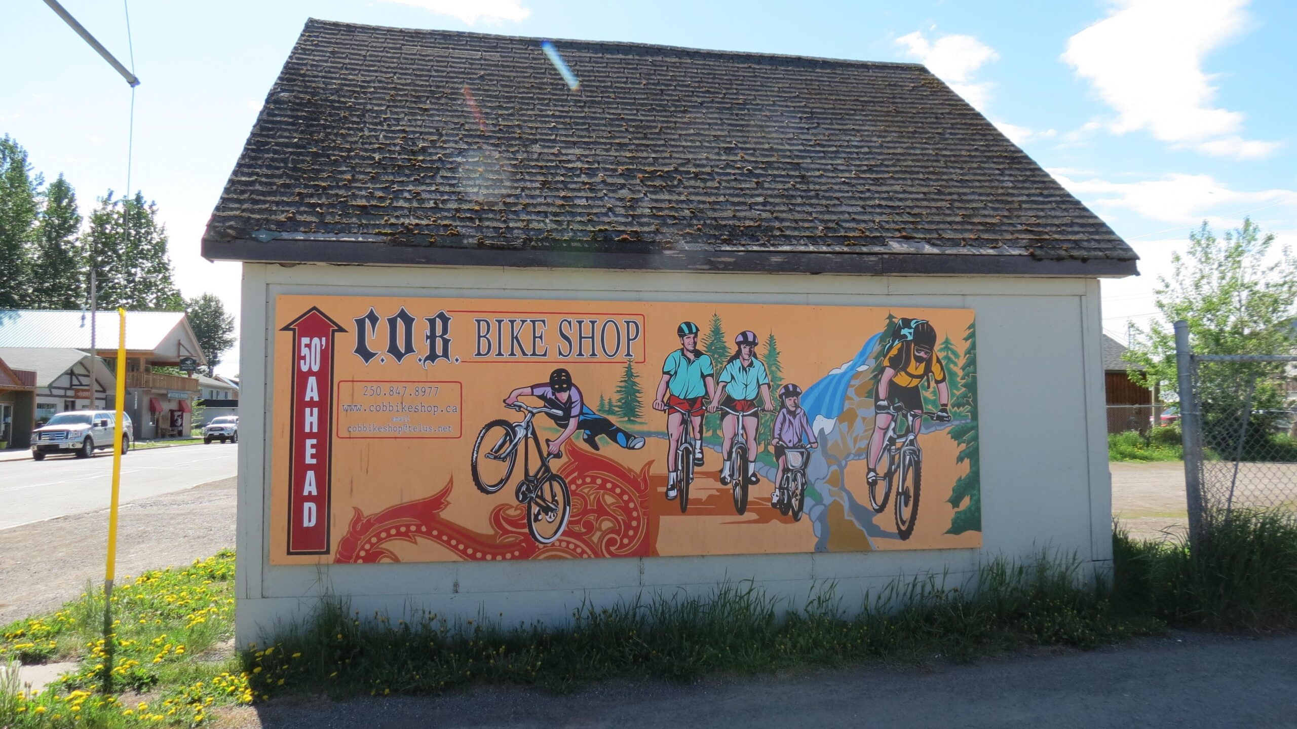 A colorful mural on the exterior of a building, promoting "C.O.B. Bike Shop." The mural features several cyclists riding bikes in a natural landscape, with mountains and trees in the background. The sign includes the shop