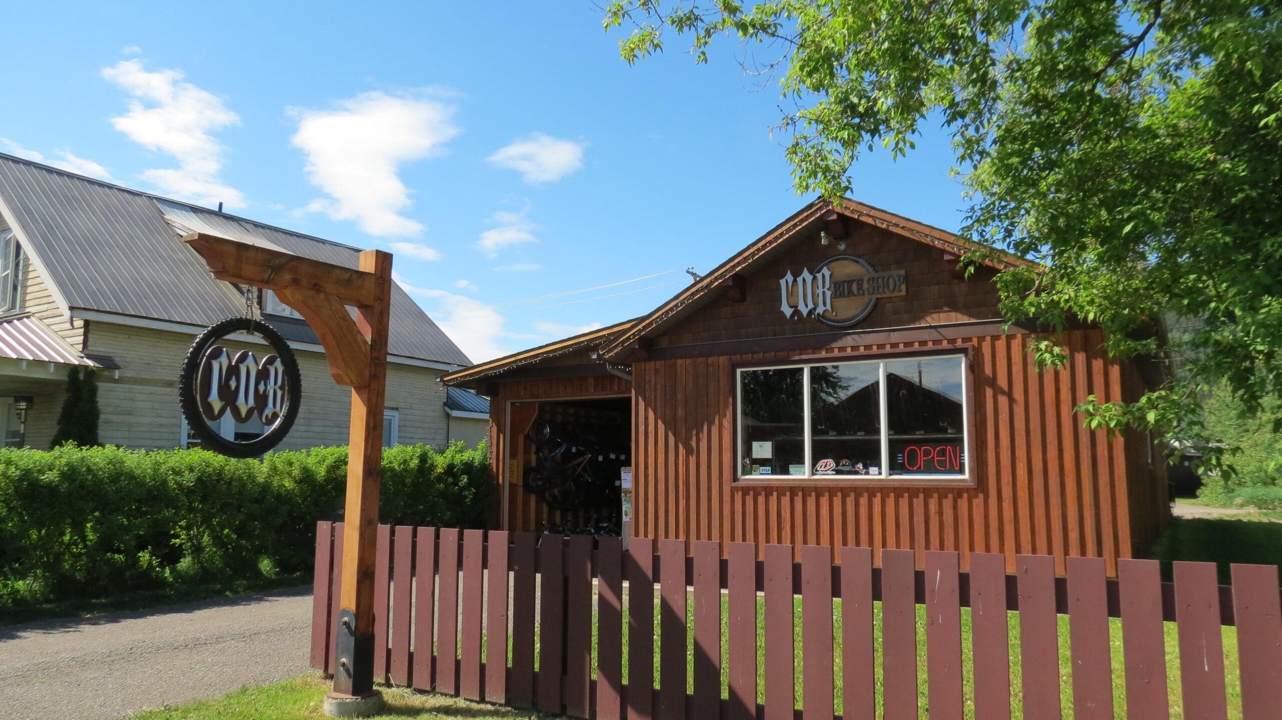 A small wooden bike shop with a sign that reads "Bike Shop" in decorative lettering. The shop has large windows displaying bicycles inside and an "OPEN" sign. A wooden fence surrounds the property, and the sky is bright with a few clouds, creating a sunny atmosphere.
