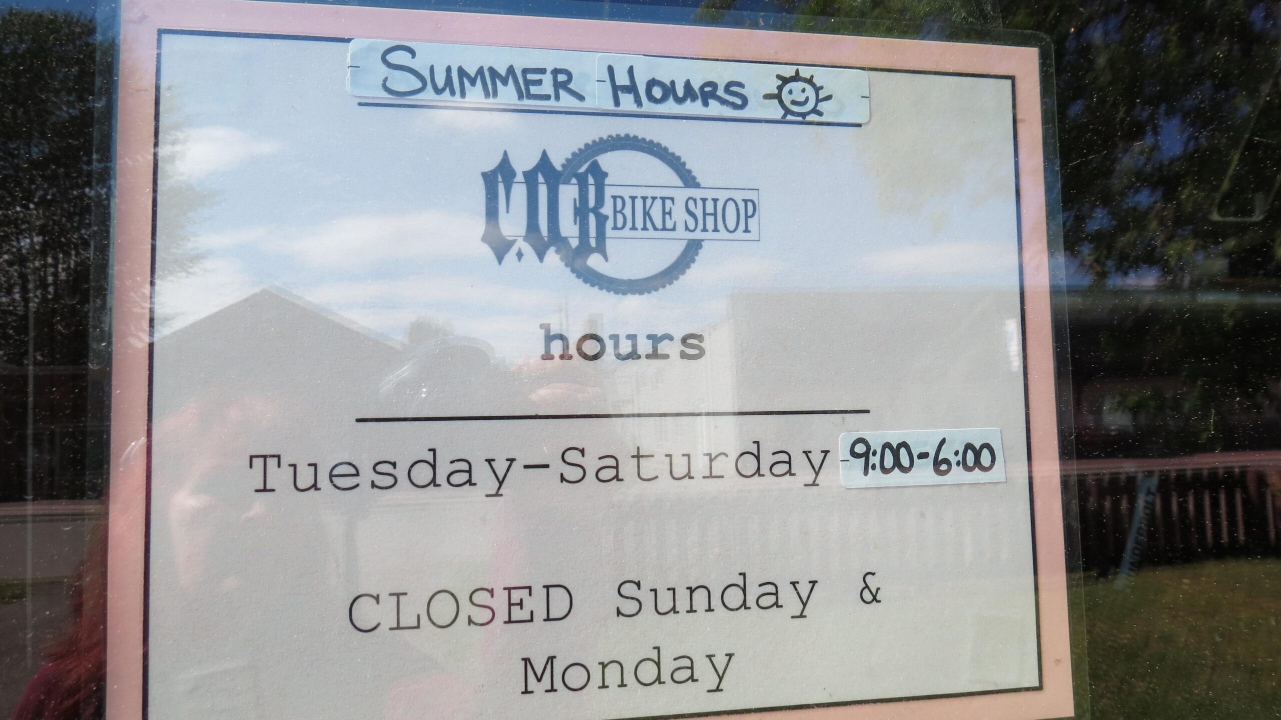 Sign on a storefront window displaying summer hours for a bike shop. The sign features "SUMMER HOURS" at the top, followed by the shop's name, "BIKE SHOP." Below, it lists the operating hours as Tuesday to Saturday from 9:00 AM to 6:00 PM, and states that the shop is closed on Sundays and Mondays.