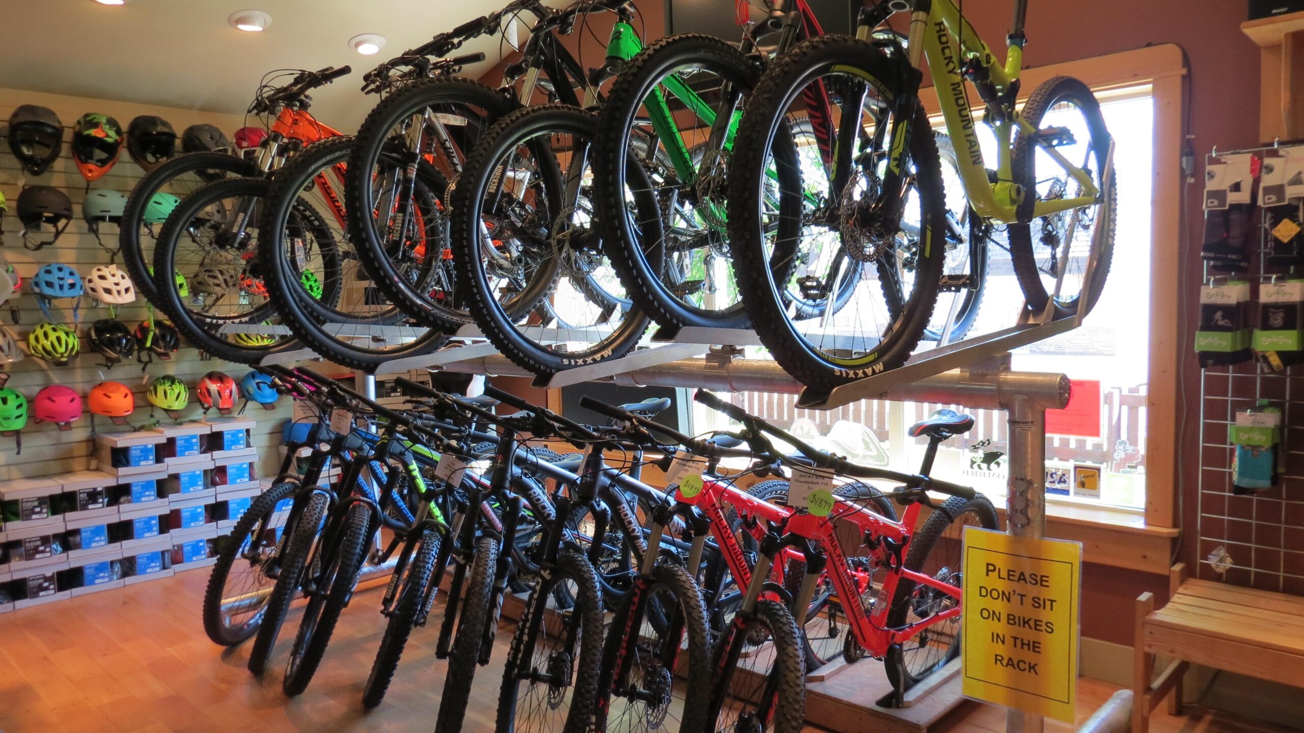 Bikes displayed in a retail shop with a variety of mountain bikes on racks and a wall of colorful helmets in the background. A sign instructs customers not to sit on the bikes.