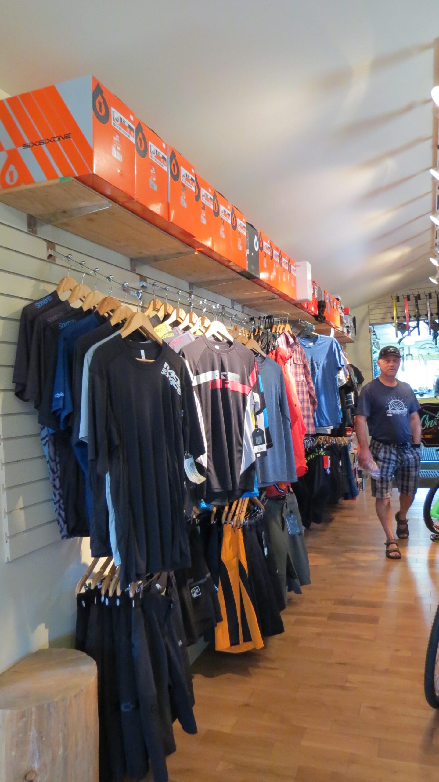 A retail shop interior featuring a display of cycling apparel on hangers, including T-shirts and shorts. Brightly colored packaging boxes are stacked on shelves above. A man in casual clothing walks through the store, with wooden flooring visible. The space is well-lit, showcasing various biking accessories and gear.