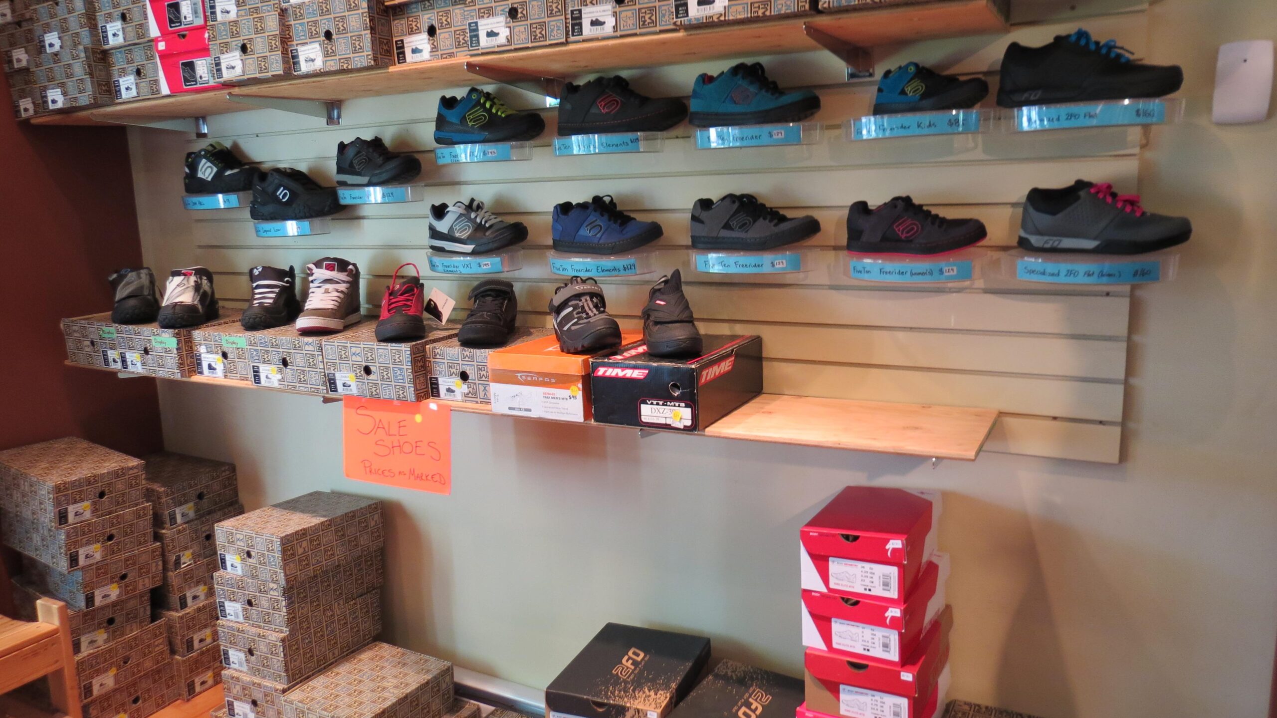 A display of various athletic shoes arranged on shelves in a store. Some shoes are hanging on plastic shelves, while others are placed on boxes below. A sign in the foreground indicates that there are shoes on sale with prices unmarked. The store has a warm interior, with shoe boxes stacked neatly in the bottom right corner.