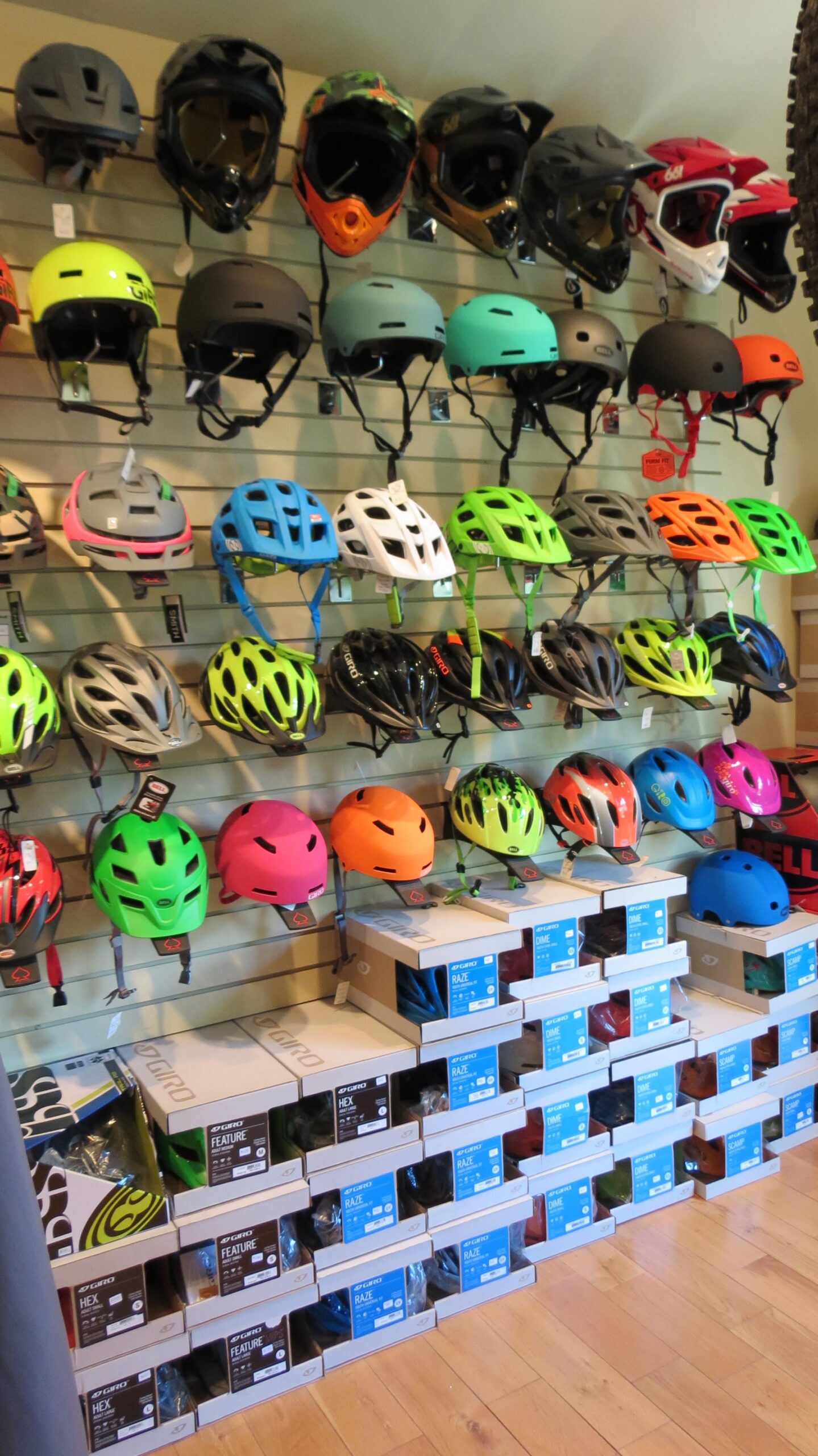 A display of various colorful helmets hanging on a wall, with additional boxes of helmets stacked on the floor below. The helmets come in different styles and colors, including bright shades of green, pink, orange, and black, showcasing a variety of brands. The wooden floor adds a warm tone to the indoor setting.