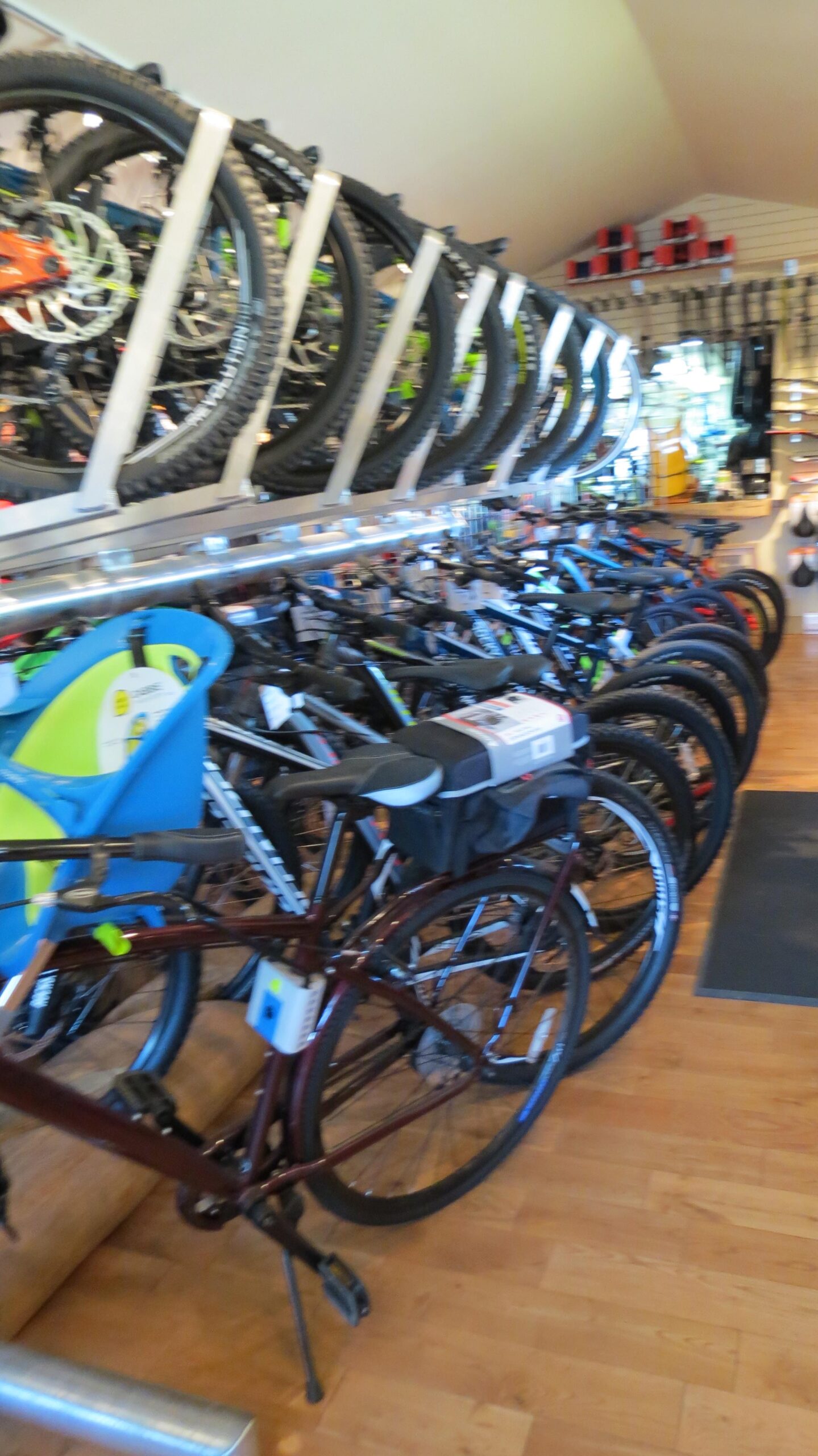 A rows of various bicycles displayed in a bike shop, featuring different styles and colors. A blue and green child seat is visible in the foreground, while other bikes with varying designs and accessories are lined up on the floor and mounted on overhead racks. The interior of the shop has wooden flooring and a bright atmosphere, with additional cycling gear visible in the background.