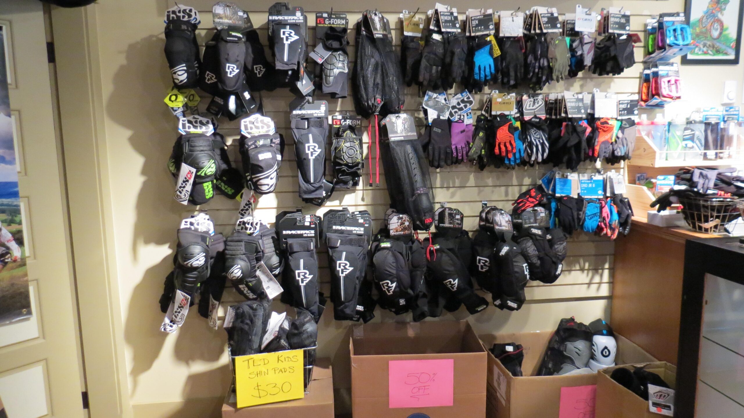 Display of various types of protective gloves and shin pads hanging on a wall in a shop. The gloves include bright colors and different styles, with some labeled for discounts. There are also boxes at the bottom indicating a sale on certain items.