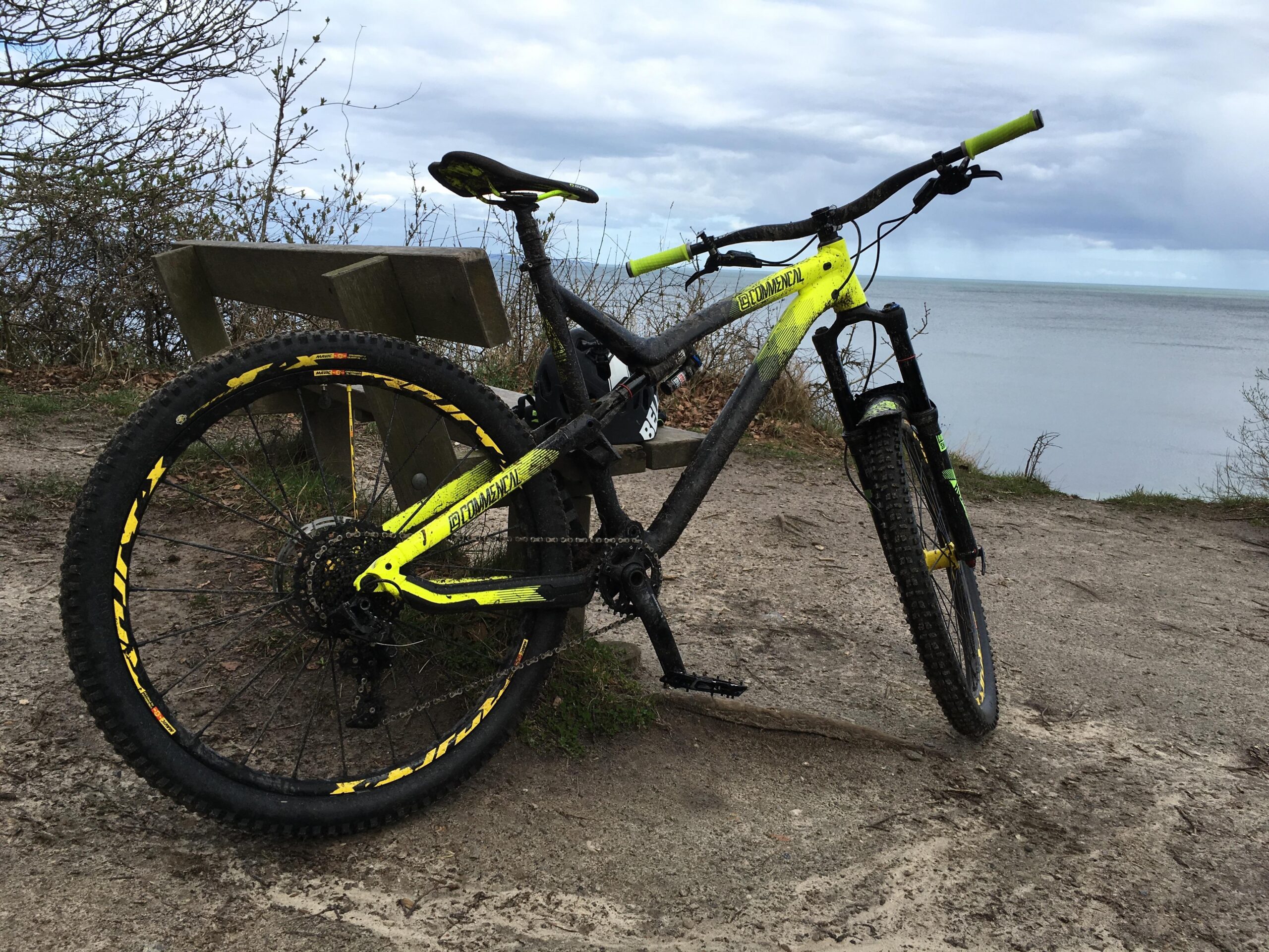 Commencal Meta AM V4: A mountain bike with a black and bright yellow frame resting on a sandy trail near a wooden bench, overlooking a calm body of water under a cloudy sky.