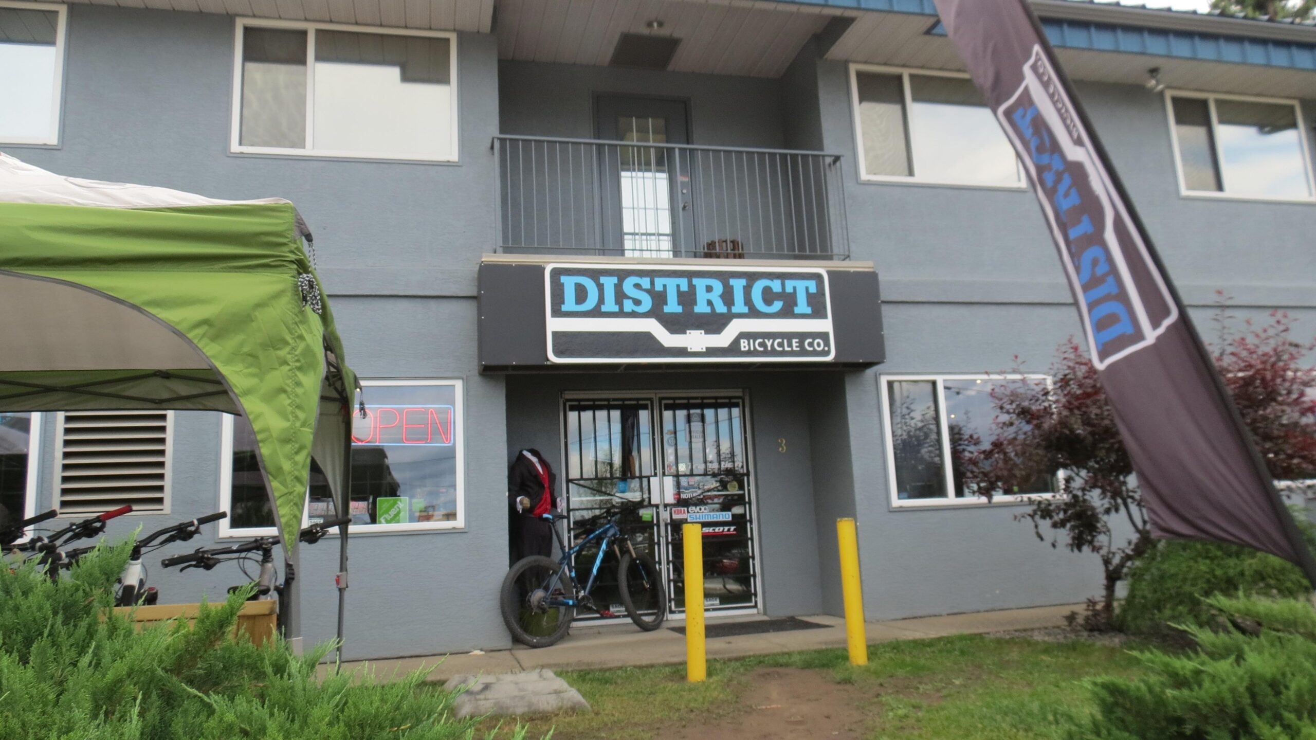 Exterior view of District Bicycle Co. showing a blue and black sign with the company's name. The entrance is adorned with a "OPEN" neon sign. A bicycle is propped against the building, and a mannequin dressed in a black jacket is next to the door. Yellow poles are positioned in front of the shop, with greenery and a banner featuring the company name visible in the surrounding area.