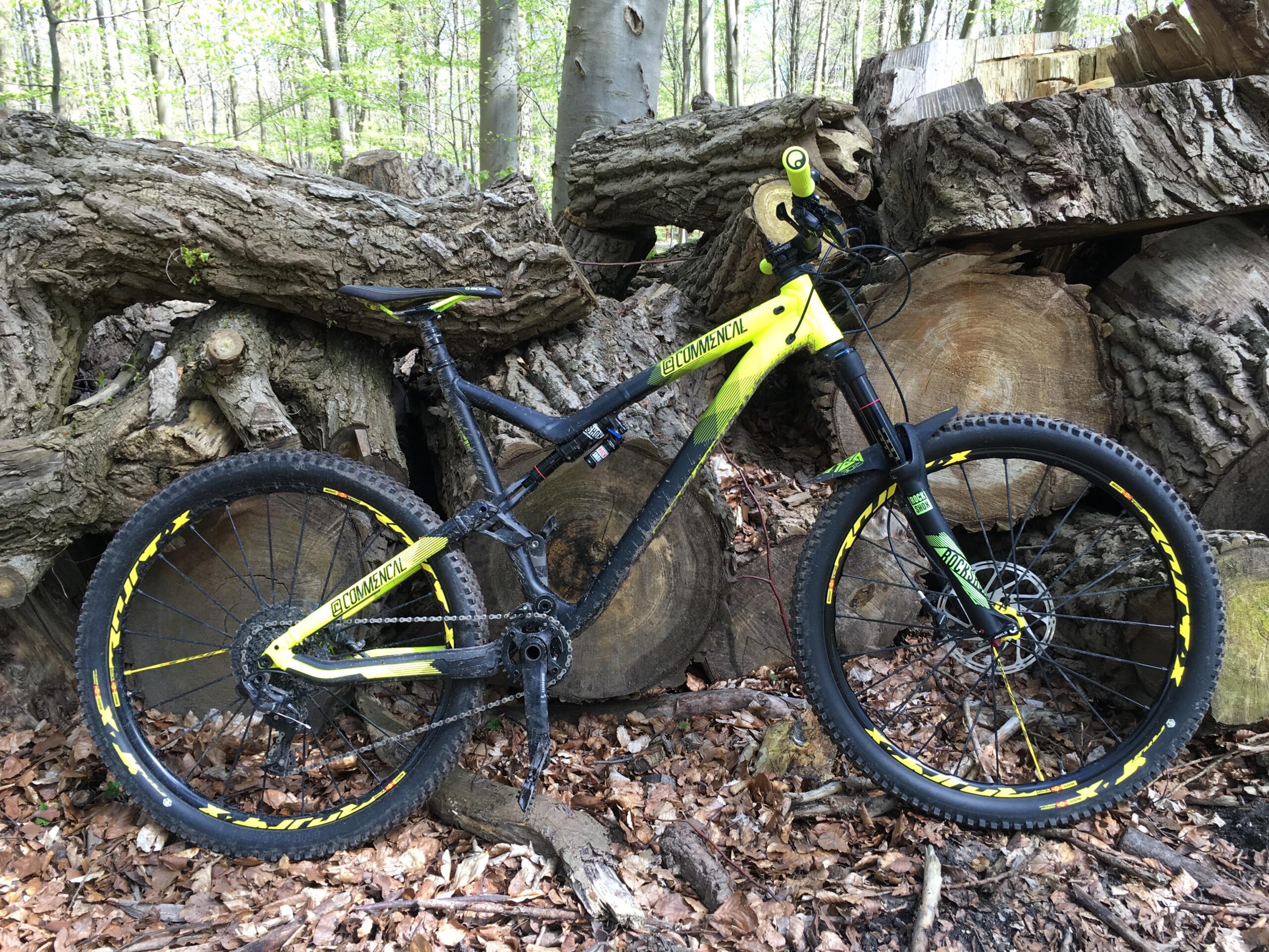 Commencal Meta AM V4: A mountain bike with a black and bright yellow frame is leaning against a stack of logs in a wooded area. The ground is covered with fallen leaves, and fresh green leaves are visible on the trees in the background. The bike features prominent tires designed for off-road terrain and has various components, including a suspension fork and disc brakes.