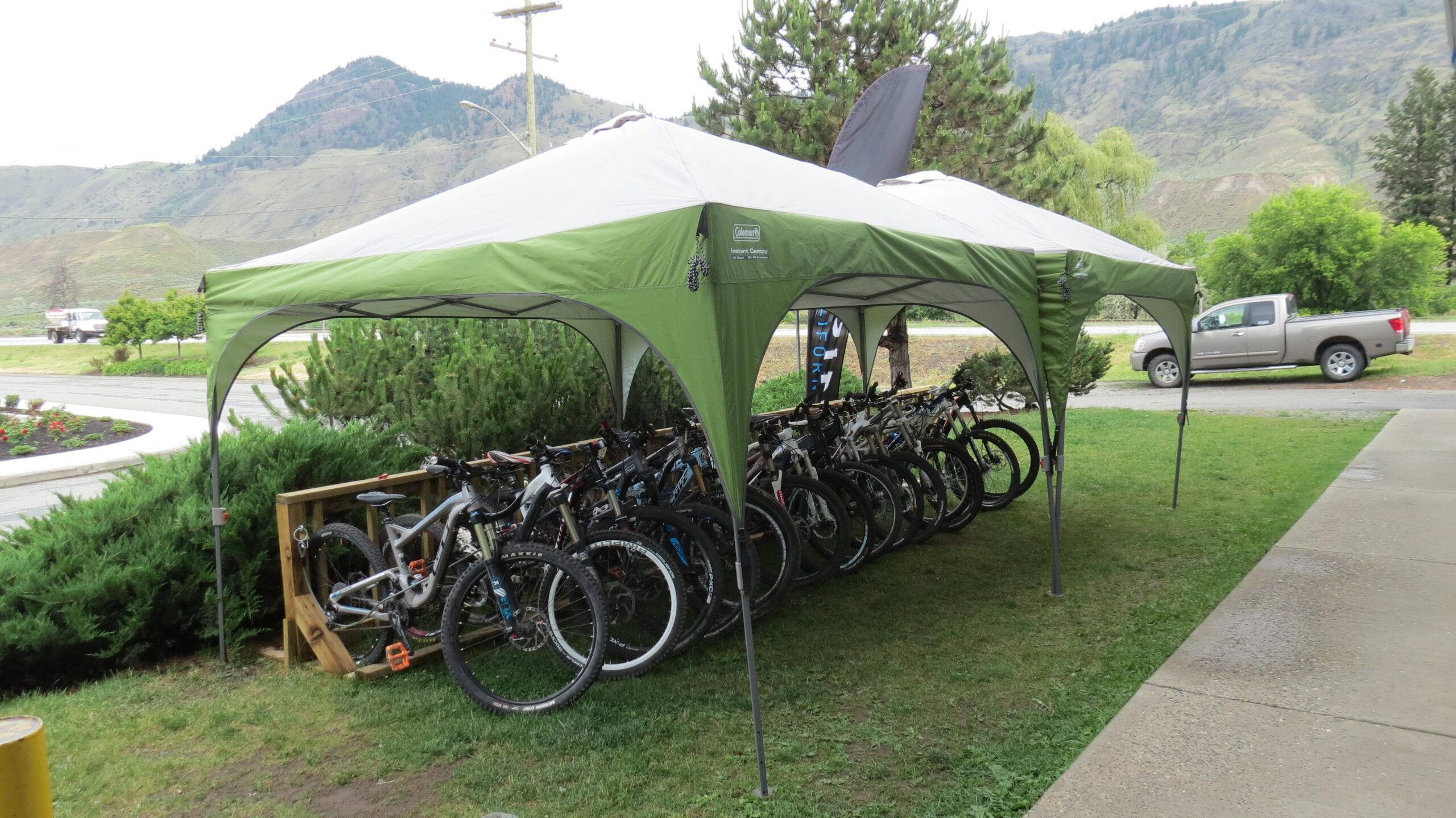 A green tent set up outdoors with a row of mountain bikes underneath. The background features hills and trees, and there’s a parked truck nearby on a paved area. The scene is in a grassy area with some shrubs and plants.