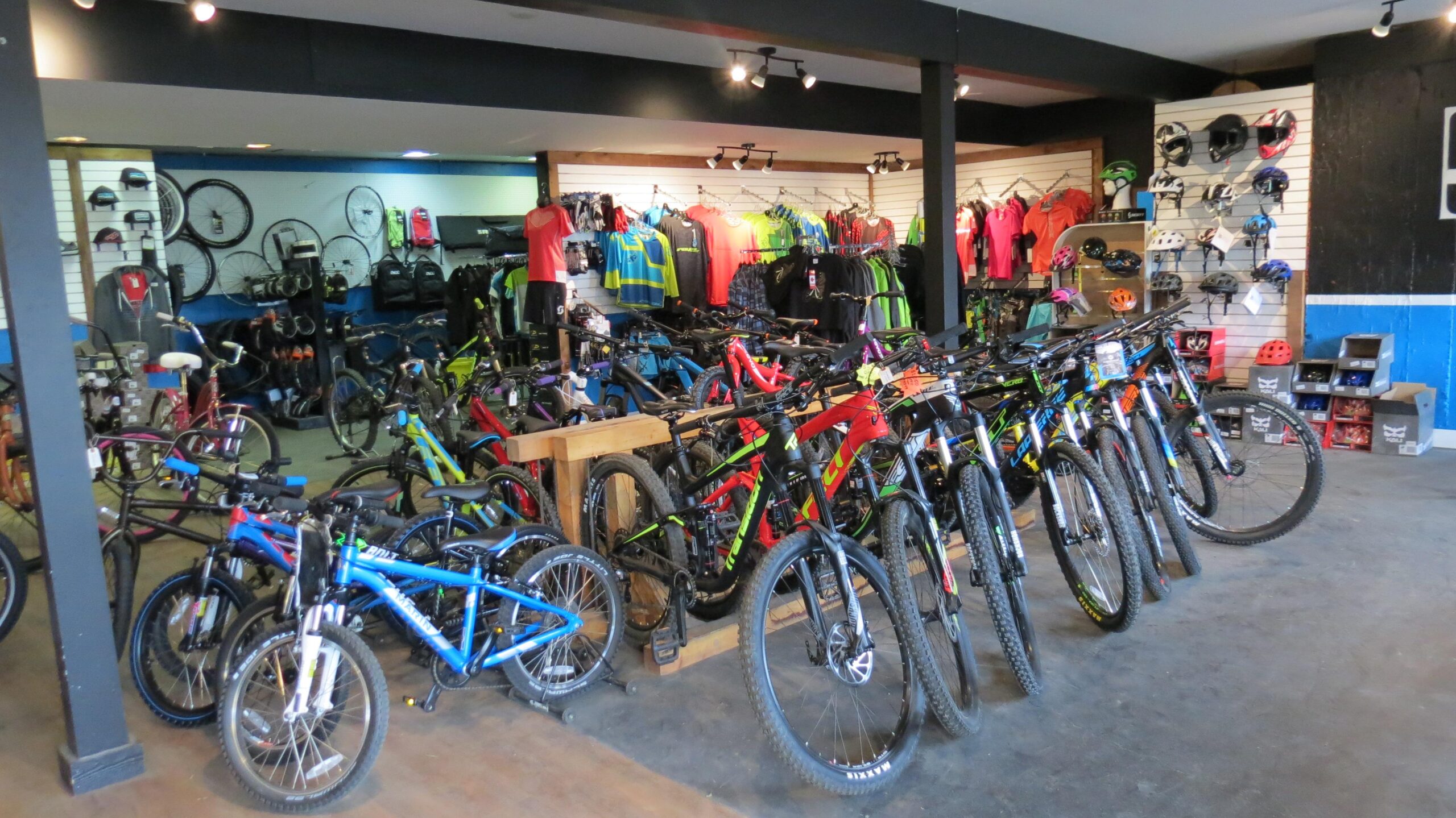 A vibrant bike shop interior featuring a variety of bicycles, including mountain bikes and kids' bikes, displayed on the floor and on wooden stands. The walls are adorned with cycling apparel, helmets, and accessories, creating an inviting atmosphere for cycling enthusiasts.
