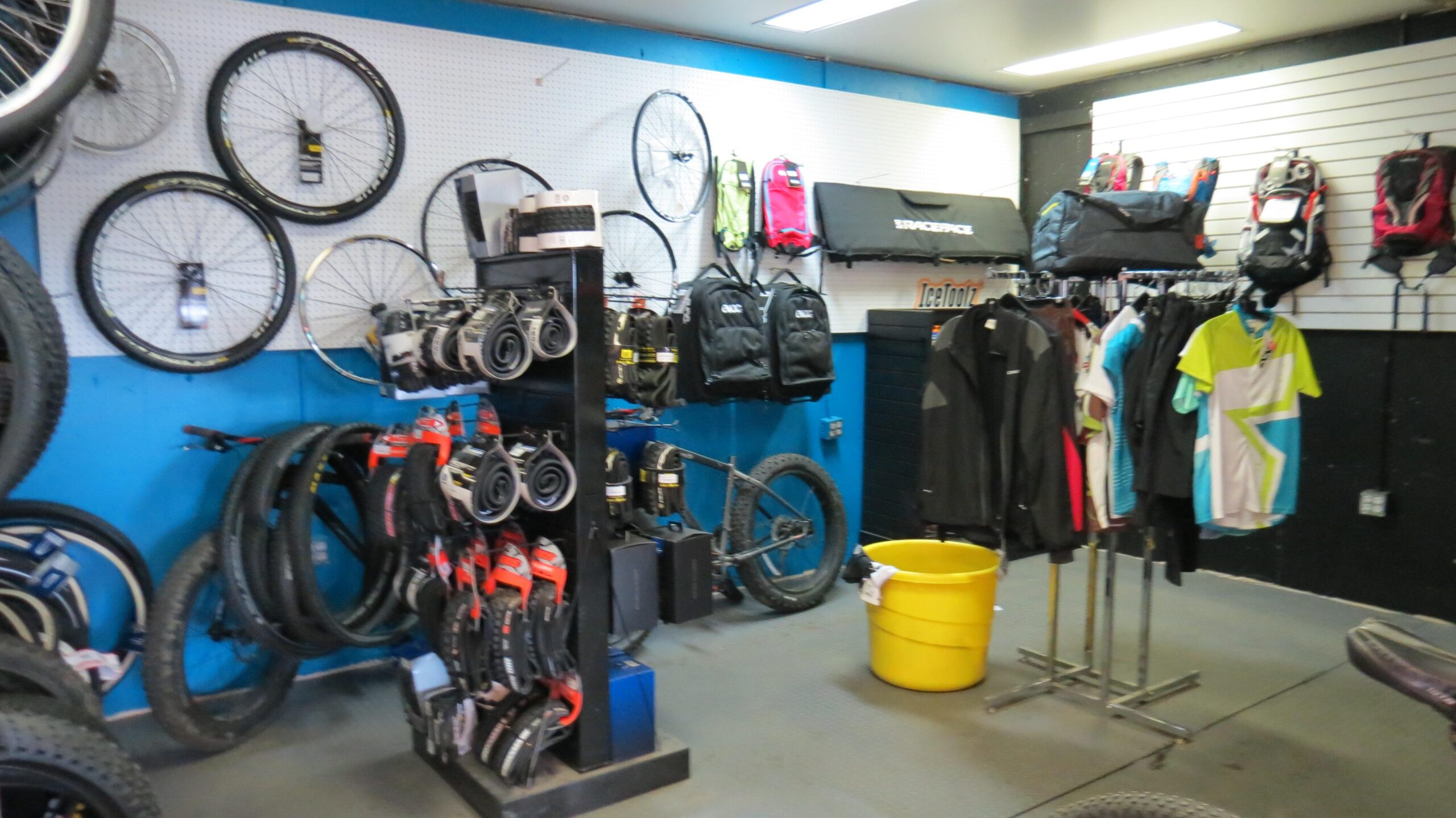 A bike shop interior featuring various bicycle wheels and tires displayed on the walls, along with racks of cycling apparel, backpacks, and gear. A yellow bucket is positioned on the floor, and a bicycle frame is partially visible among the displayed items. The walls are painted blue and black, creating a vibrant shopping atmosphere for cycling enthusiasts.
