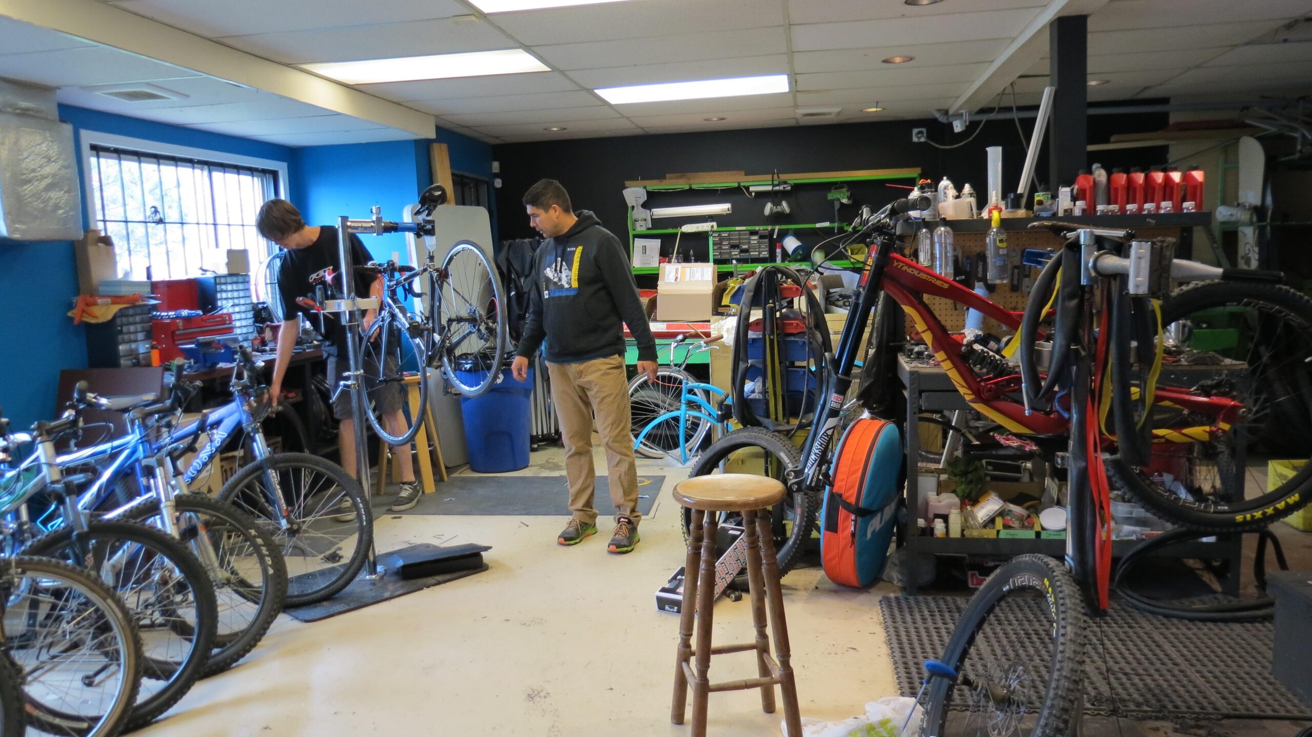 A cluttered bicycle workshop with two individuals working on bicycles. One person is adjusting a bike on a stand, while the other is standing nearby, looking at a different bike. Various types of bikes, tools, and repair equipment are visible throughout the space, which has blue and black walls and good lighting. A wooden stool is positioned in the foreground.