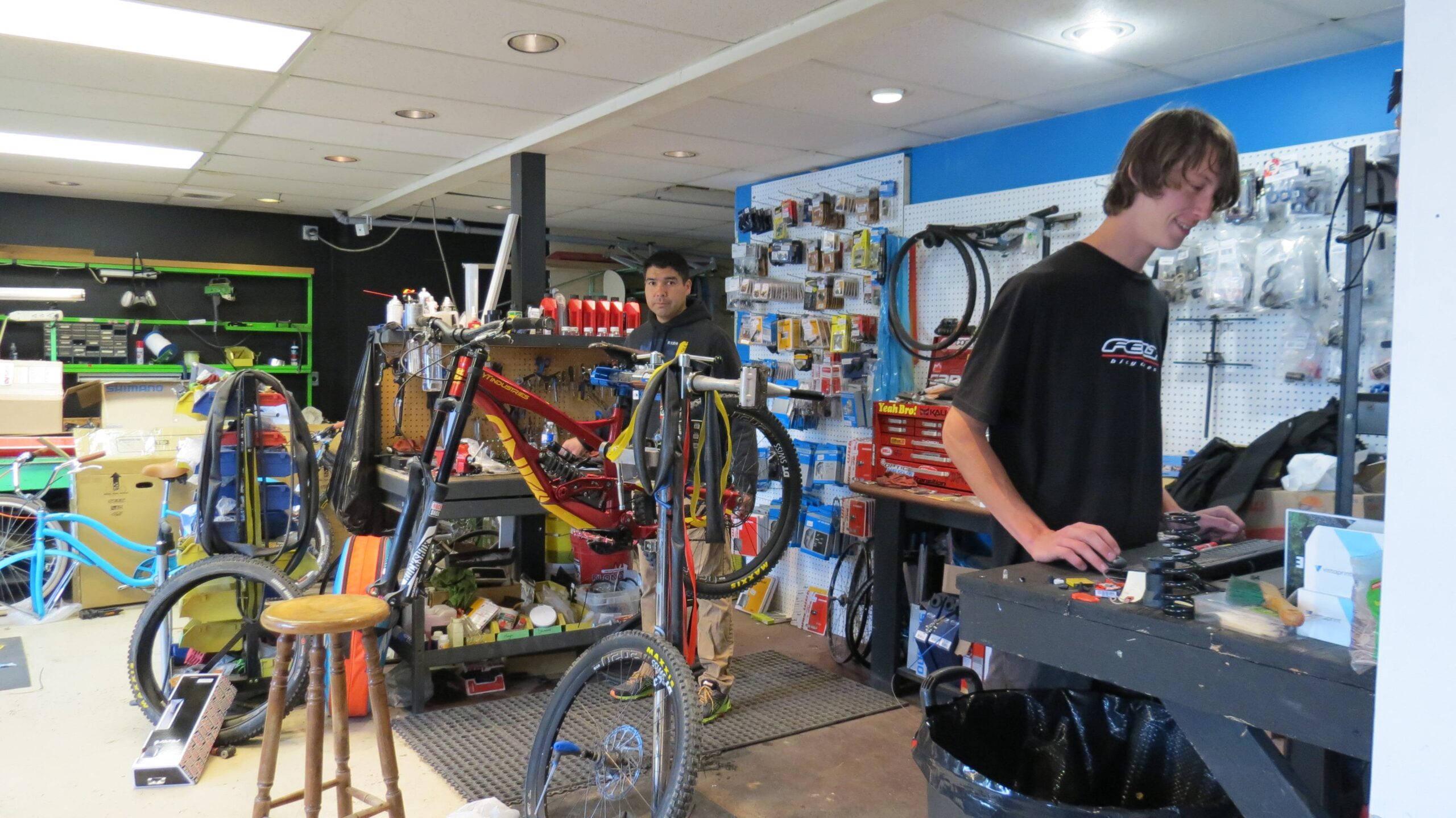 Two individuals work in a busy bike repair shop. One person, standing at a workbench with tools and bike parts, types on a laptop, while the other, positioned behind a bike held on a repair stand, looks directly at the camera. The shop is filled with various bicycles, tools, and parts displayed on shelves and work surfaces, giving it a lively workshop atmosphere.