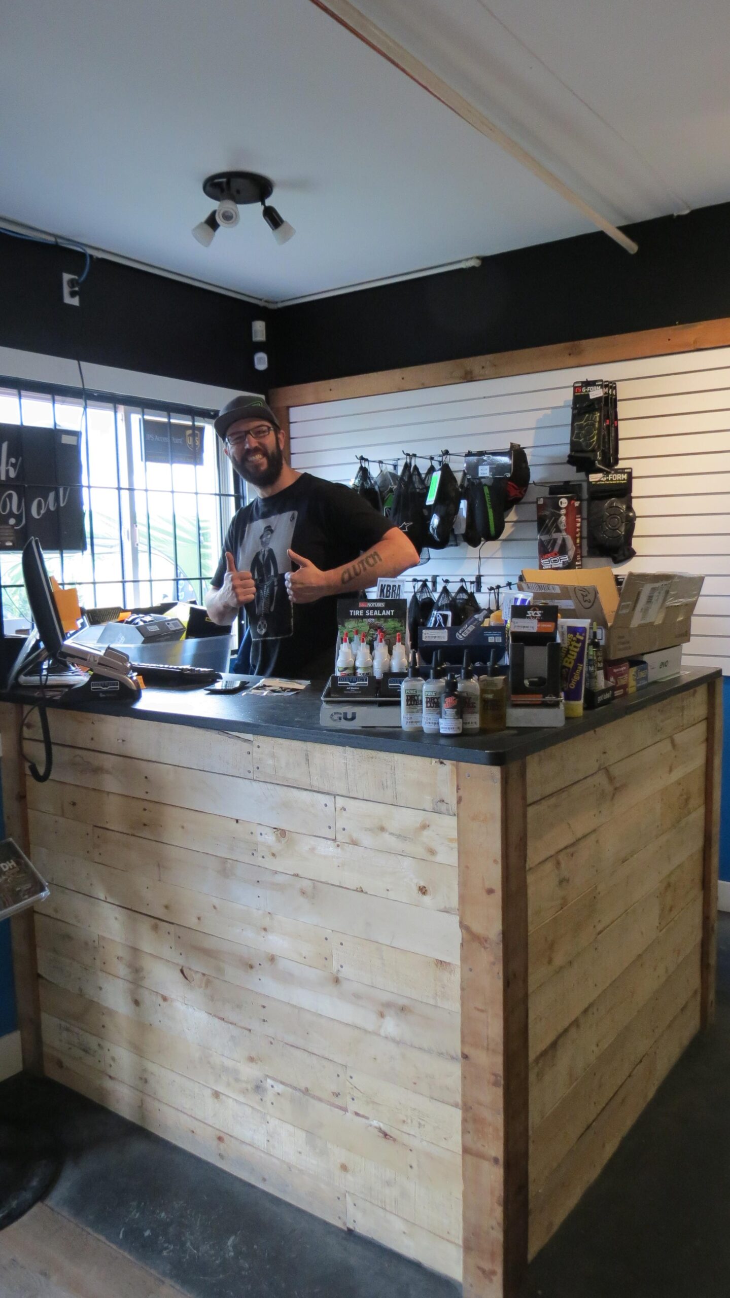A smiling employee stands behind a wooden counter in a shop, giving a thumbs-up. The store features various products displayed on shelves, including tire sealant and other bike-related items. The walls are painted in contrasting colors, with a bright area behind the counter and darker tones above.