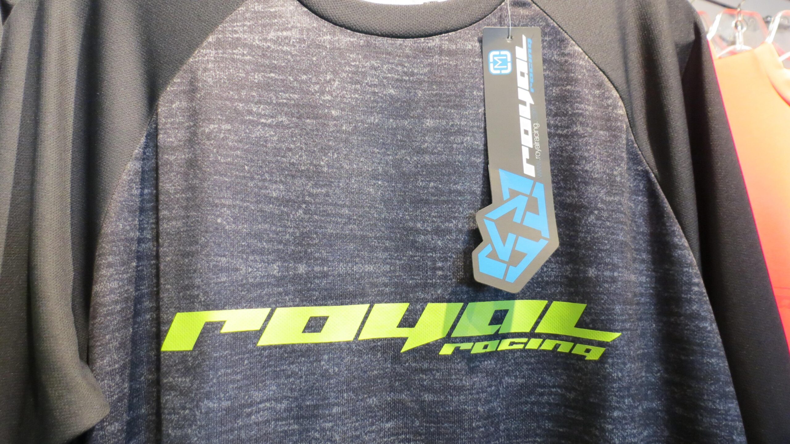 A close-up image of a black and gray athletic shirt featuring the logo "royal racing" in bright green lettering. The shirt has a textured fabric design and a tag attached, displaying the brand logo.