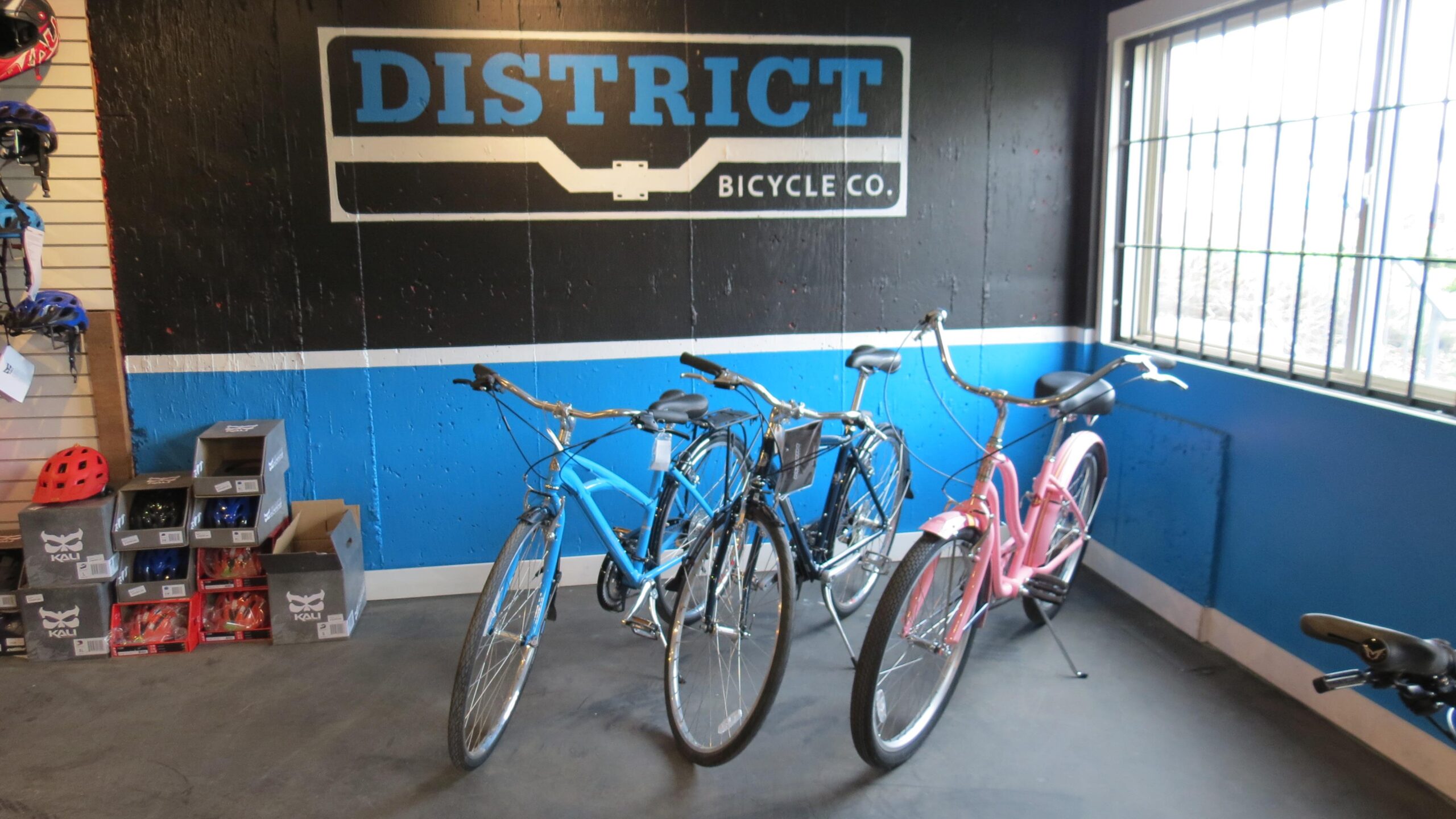 A display of bicycles in a retail shop featuring three bikes—one blue, one silver, and one pink—positioned against a black and blue wall with the words "DISTRICT BICYCLE CO." visible. Helmets and bicycle accessories are arranged on shelves in the background. Natural light shines through a nearby window.