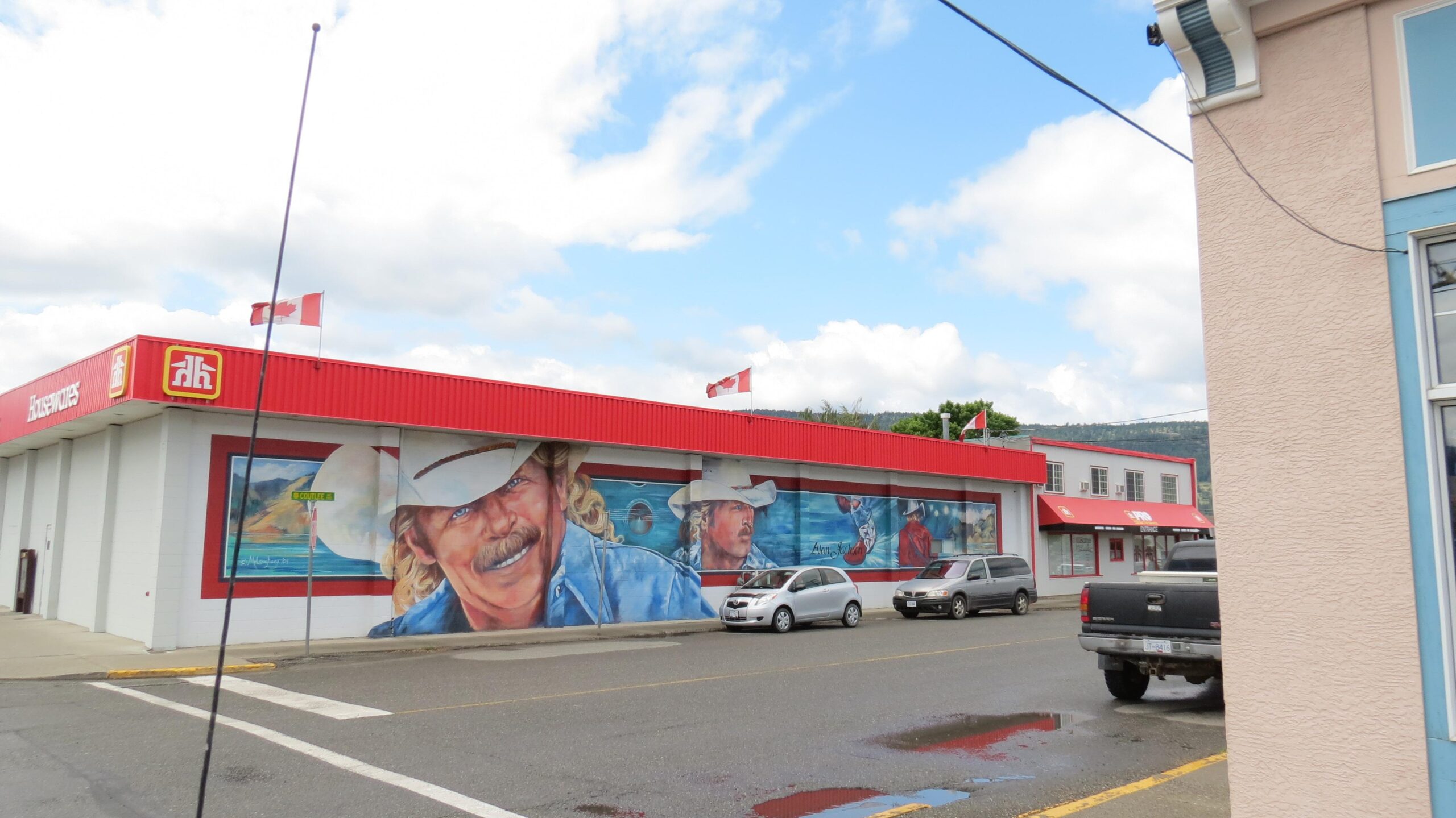 A street view of a building featuring a large mural on its side, depicting two individuals with cowboy hats and distinct facial expressions. The building is painted in red and white, with Canadian flags flying from the roof. Several cars are parked along the street, and the sky is partly cloudy.