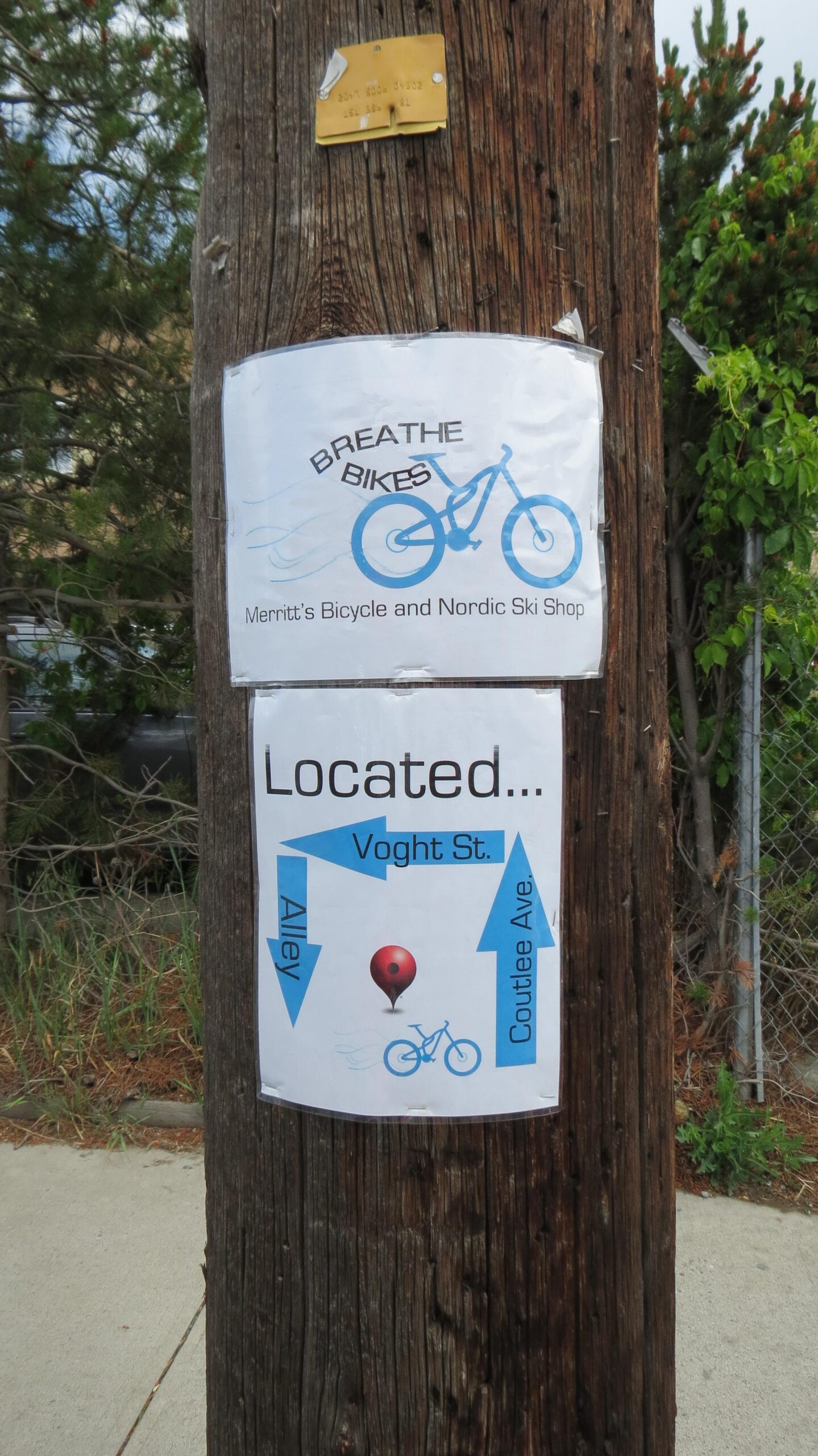A telephone pole displaying two signs. The upper sign features the text "Breathe Bikes" along with a graphic of a bicycle, advertising Merritt's Bicycle and Nordic Ski Shop. The lower sign provides directional information, indicating the location as "Located... Voght St." with arrows pointing to "Alley" and "Coutlee Ave." along with a red balloon graphic.