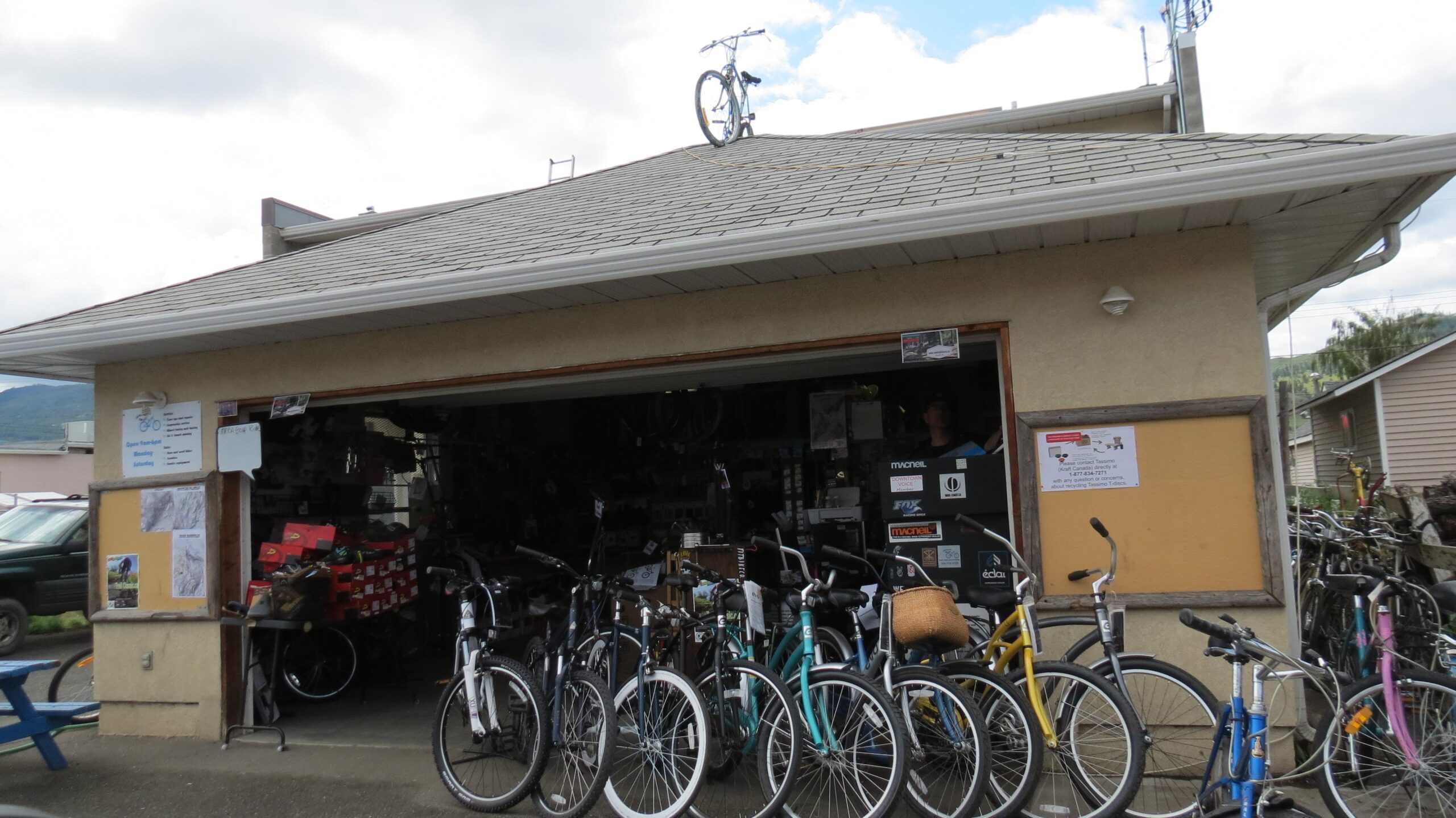 A bicycle shop with various bicycles parked outside, showcasing a range of colors and styles. The shop has an open entrance with a cluttered display of bike accessories visible inside. A bike is humorously perched on the roof, adding a quirky touch to the establishment. The sky is partly cloudy, with a few trees and vehicles visible in the background.