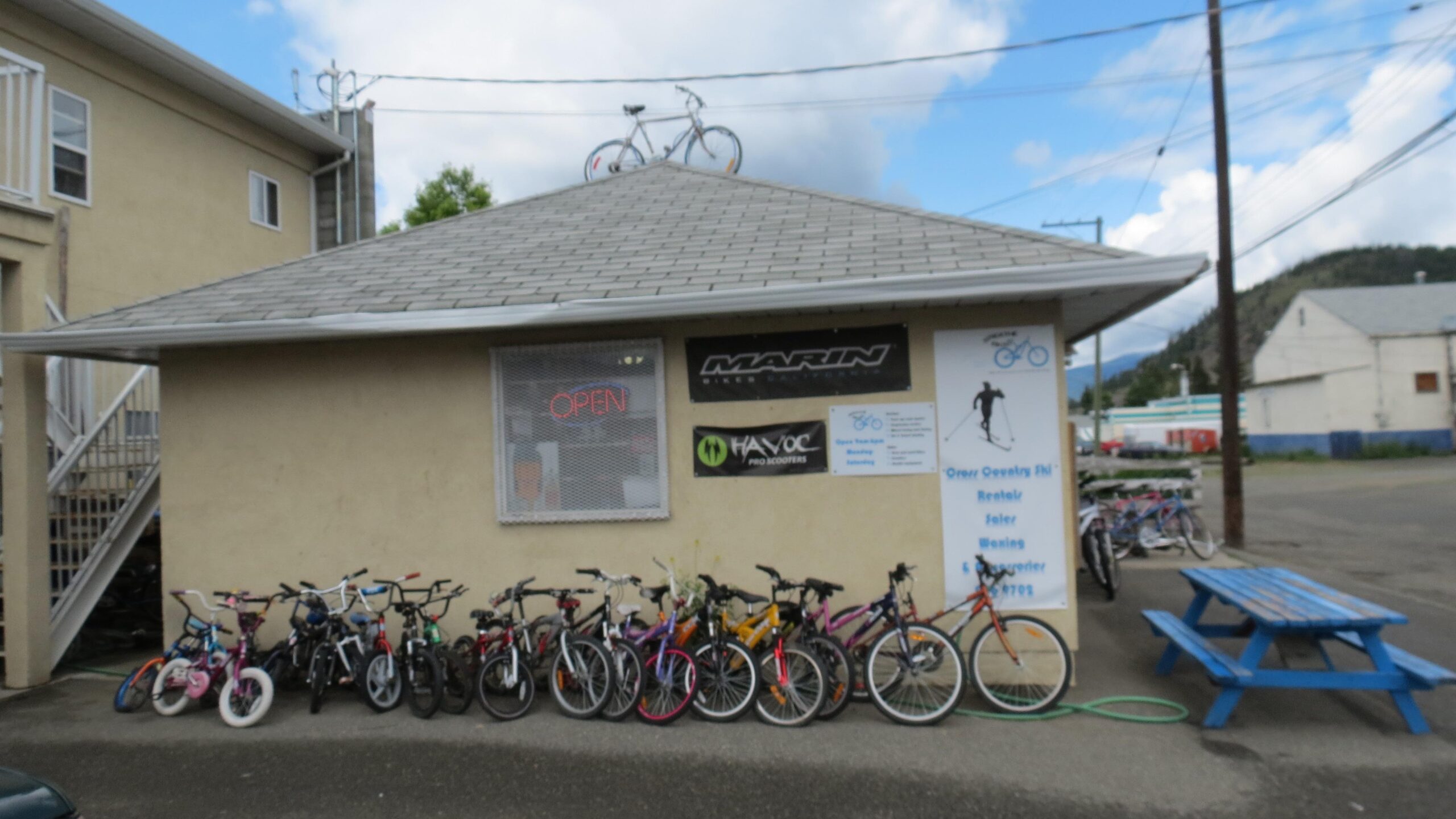 A small bike shop with a sign that says "Open," featuring an assortment of bicycles parked in front. The building has promotional banners for Marin and Havoc, and a bicycle mounted on the roof. A blue picnic table is visible on the side, with a backdrop of a cloudy sky and surrounding buildings.