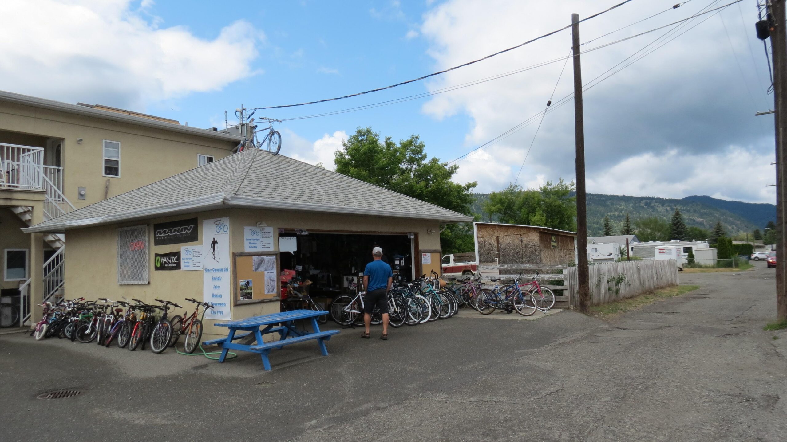 A small bike shop with a gray roof displaying a variety of bicycles in front. A person in a blue shirt is standing in front of the shop, which has an "Open" sign, with posters and information visible on its walls. The background features a scenic view of hills, trees, and a cloudy sky. There are power lines and a parking area nearby, along with other buildings in the vicinity.