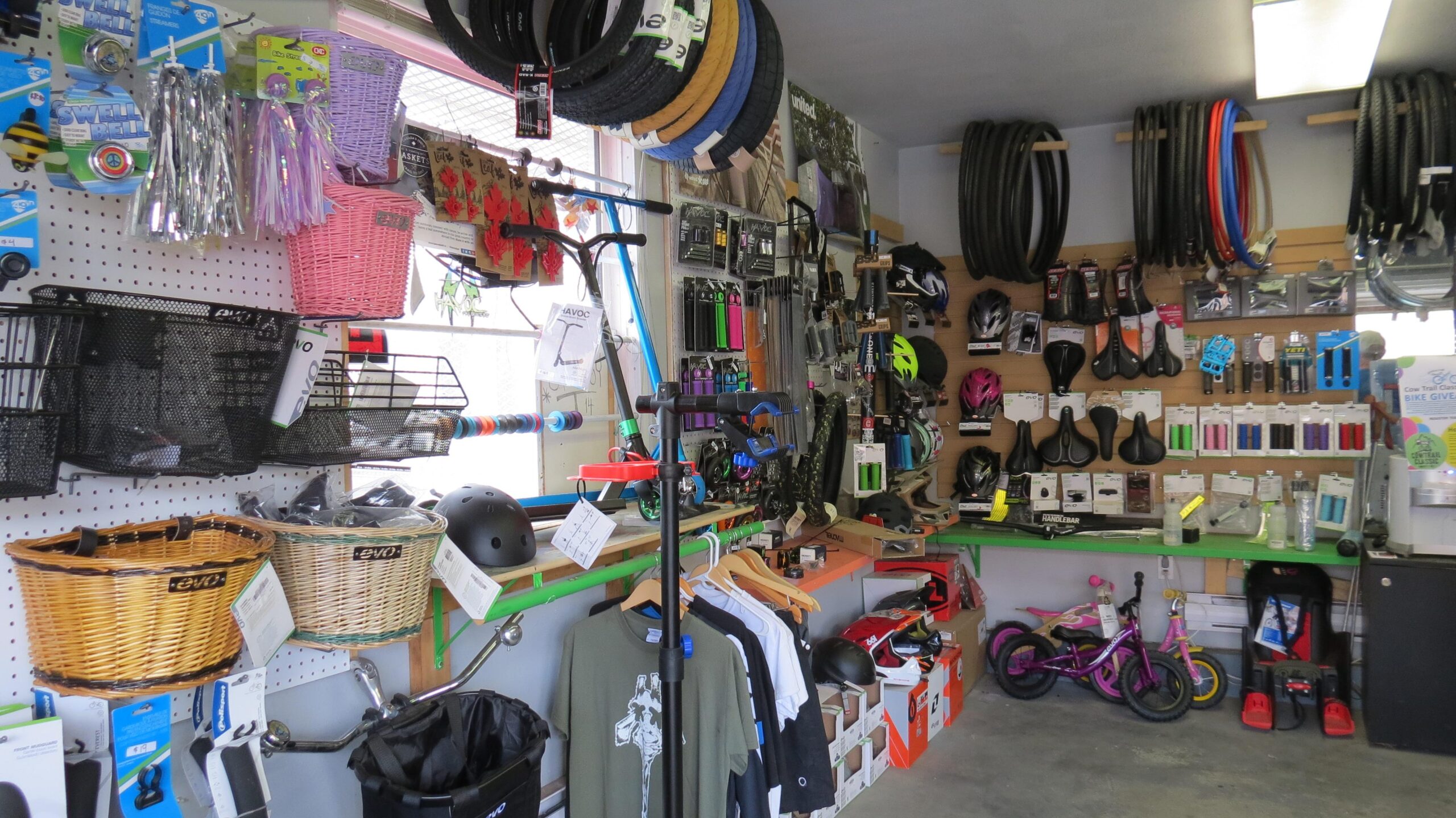 A well-organized shop interior displaying a variety of biking accessories and gear. The walls are lined with helmets, tires, bike seats, and colorful baskets. Also featured are scooters, clothing items, and various tools, creating a vibrant atmosphere for cycling enthusiasts. The setup includes shelves, pegboards, and hanging items, enhancing accessibility and visibility of the products.