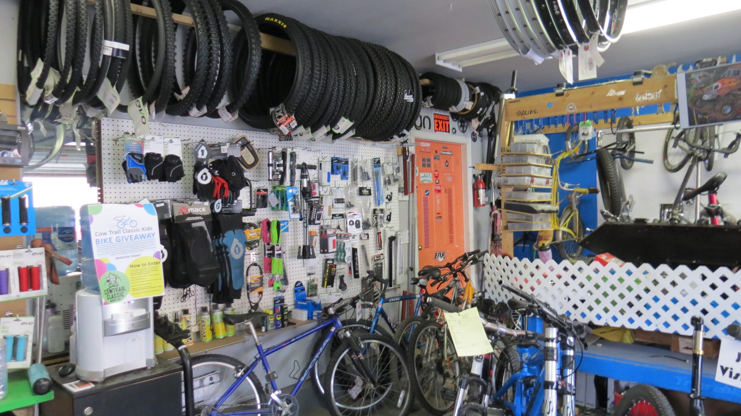 A cluttered bike shop interior featuring various bicycle tires hanging on the wall, an assortment of bike accessories displayed on pegboards, and multiple bicycles parked on the floor. A sign for a "Cow Trail Classic Kids Bike Giveaway" is visible near a water cooler. The shop also contains shelves with tools and equipment for bike maintenance.