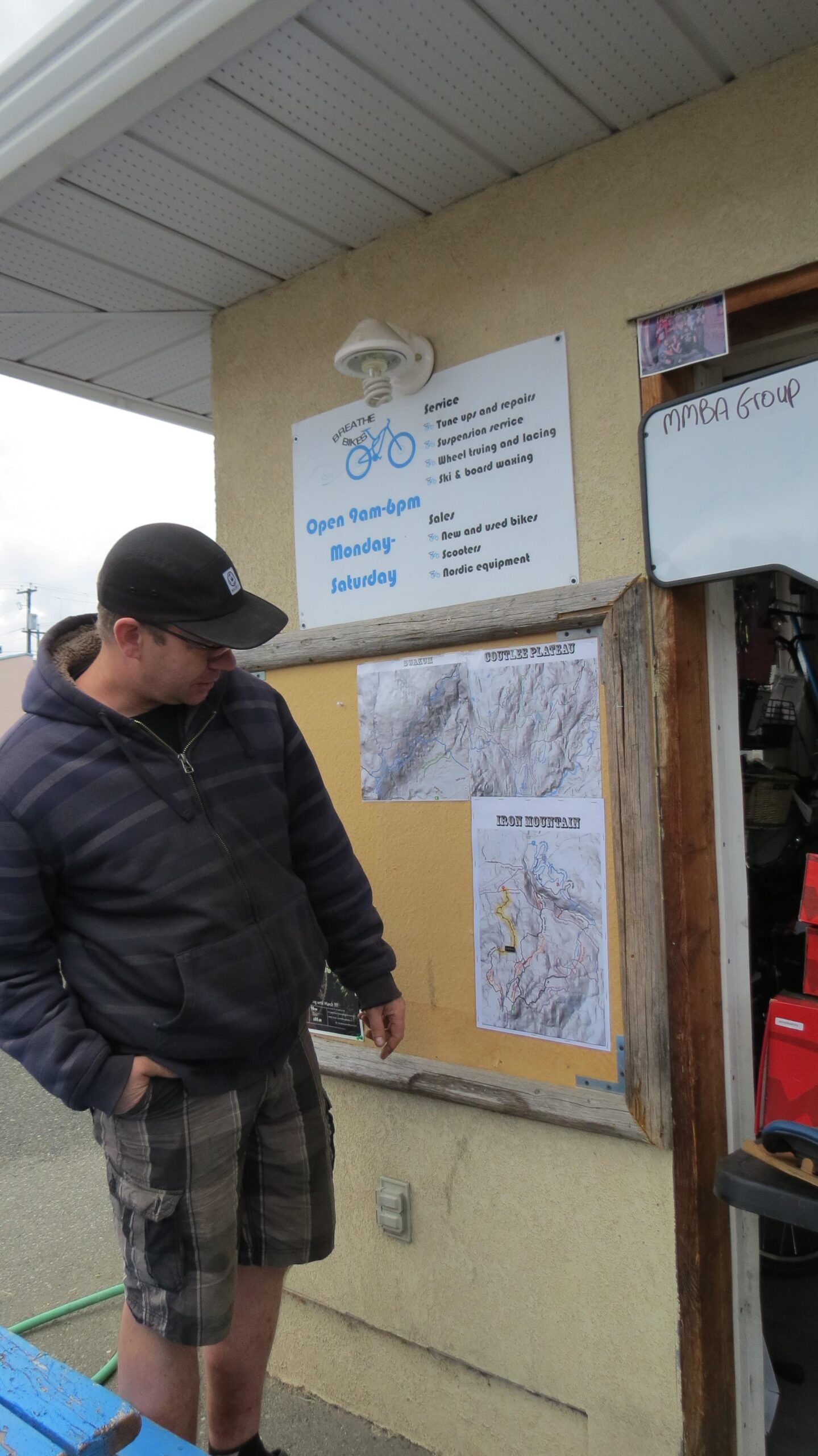 A man in a dark hoodie and shorts stands in front of a bulletin board, looking at maps labeled "South Plateau" and "Iron Mountain." Above him is a sign for a bicycle shop, detailing services and business hours. The setting appears to be outdoors, near the shop entrance, with a casual atmosphere.