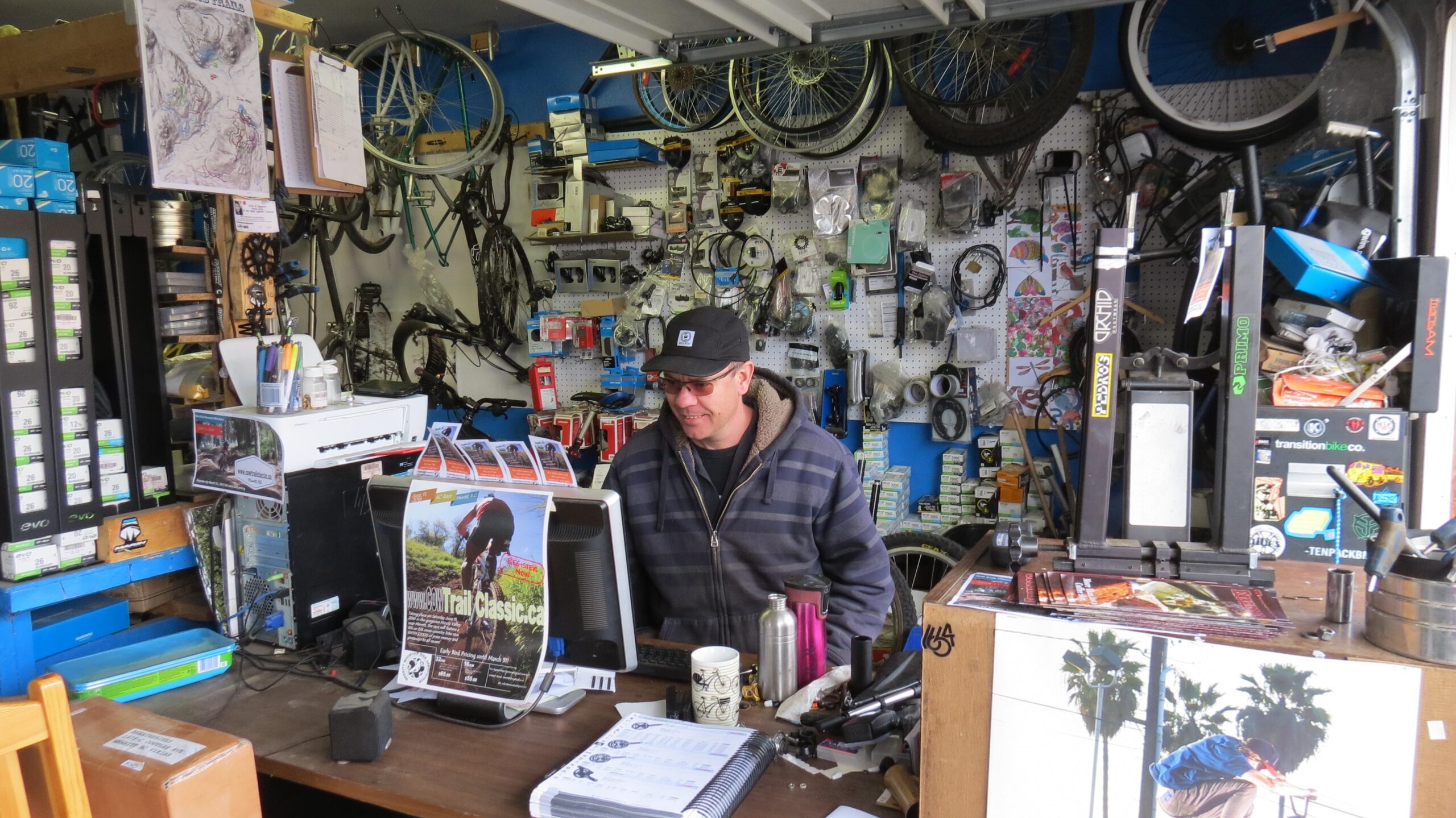 A man sitting at a desk in a bicycle shop, surrounded by various bicycle parts and accessories hanging on the walls. The desk features a computer monitor displaying a cycling event poster, and there are also cups and stationery items visible. Bicycles and bike wheels are hanging from the ceiling and walls, creating a cluttered yet vibrant atmosphere typical of a local bike shop.