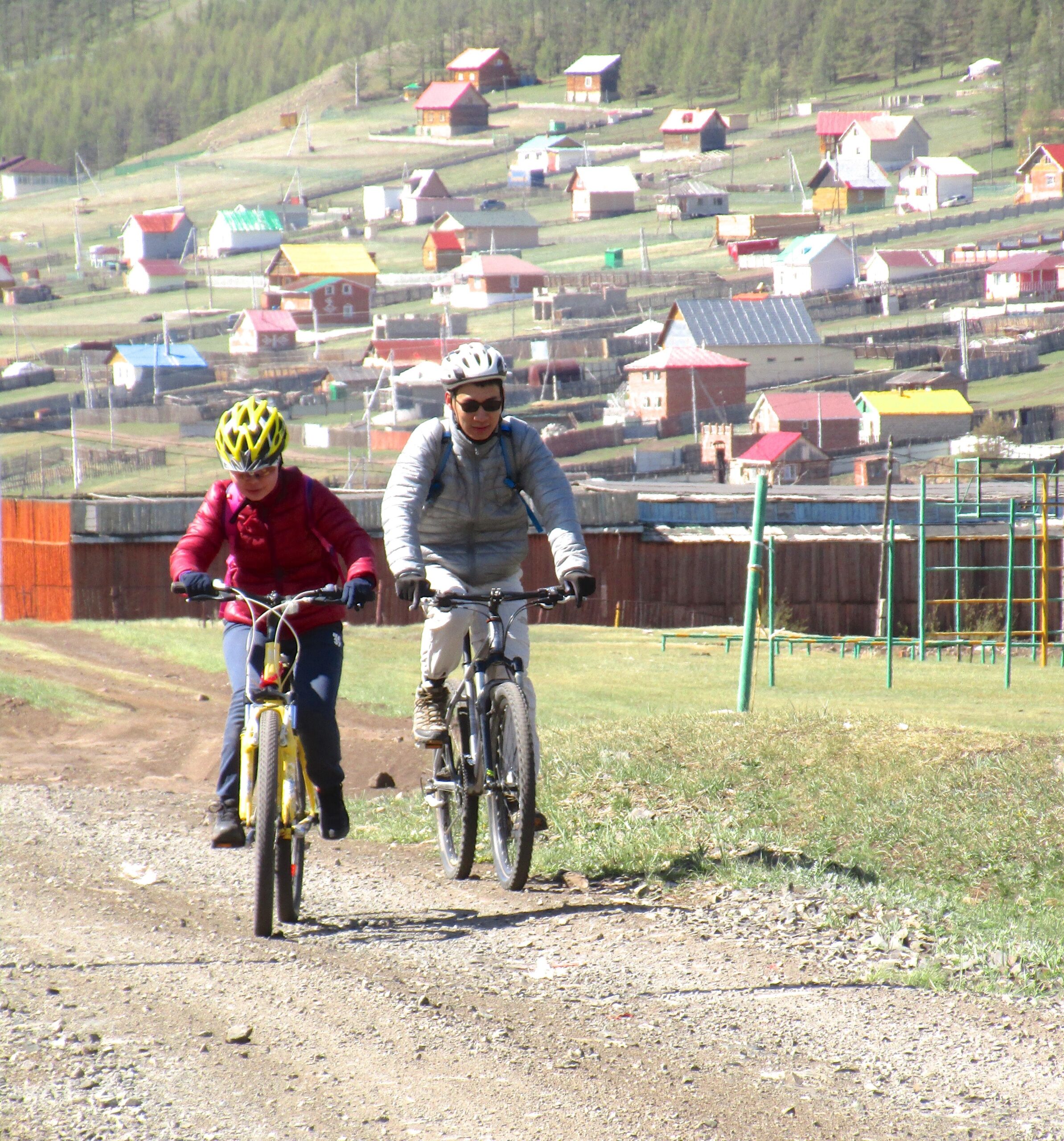 A man and a woman ride bicycles along a gravel path, surrounded by a scenic landscape featuring traditional multi-colored houses in the background. The woman wears a red jacket and a yellow helmet, while the man is dressed in a gray jacket with a helmet. Lush green grass and a playground are visible, with trees and hills in the distance. Shargamorit Khandgait Hills East mountain bike trail.