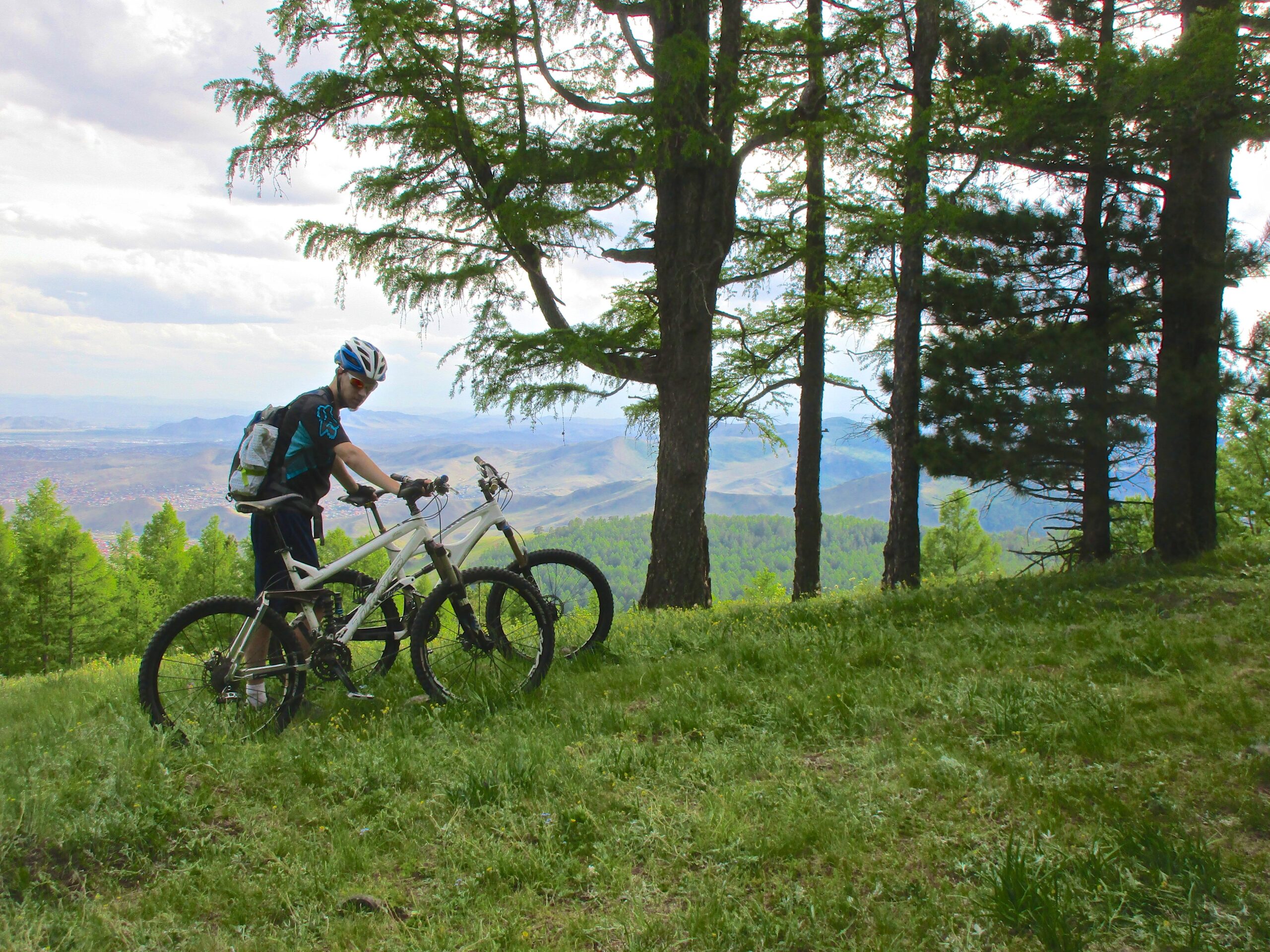 A mountain biker stands beside two bicycles in a lush green forest, surrounded by tall trees and a scenic mountainous landscape in the background. The sky is partly cloudy, and the biker is wearing a helmet and a backpack, appearing to take a moment to enjoy the view. Chengeltei Mountain North mountain bike trail.