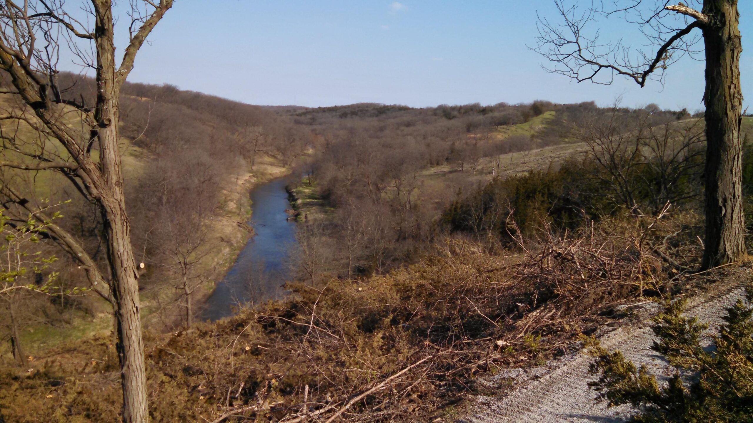 A panoramic view of a serene river winding through a hilly landscape, featuring bare trees and shrubs in early spring. The scene showcases a clear blue sky and soft sunlight illuminating the rolling terrain, with patches of grass and water visible along the riverbank. Redrock Climb- Uphill Only mountain bike trail.