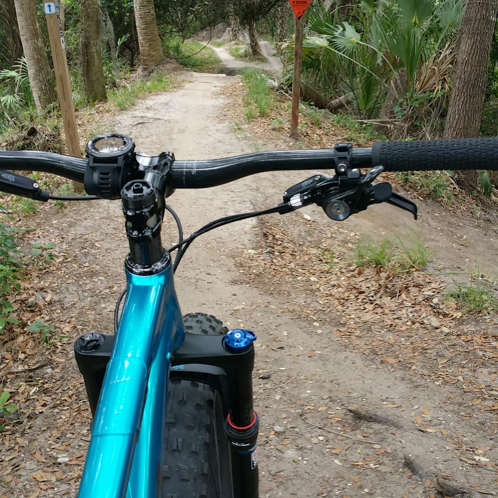 View from the handlebars of a mountain bike on a dirt trail, surrounded by trees and vegetation. The trail is winding, leading further into the forest, with a warning sign visible in the background. Grapefruit Trail mountain bike trail.