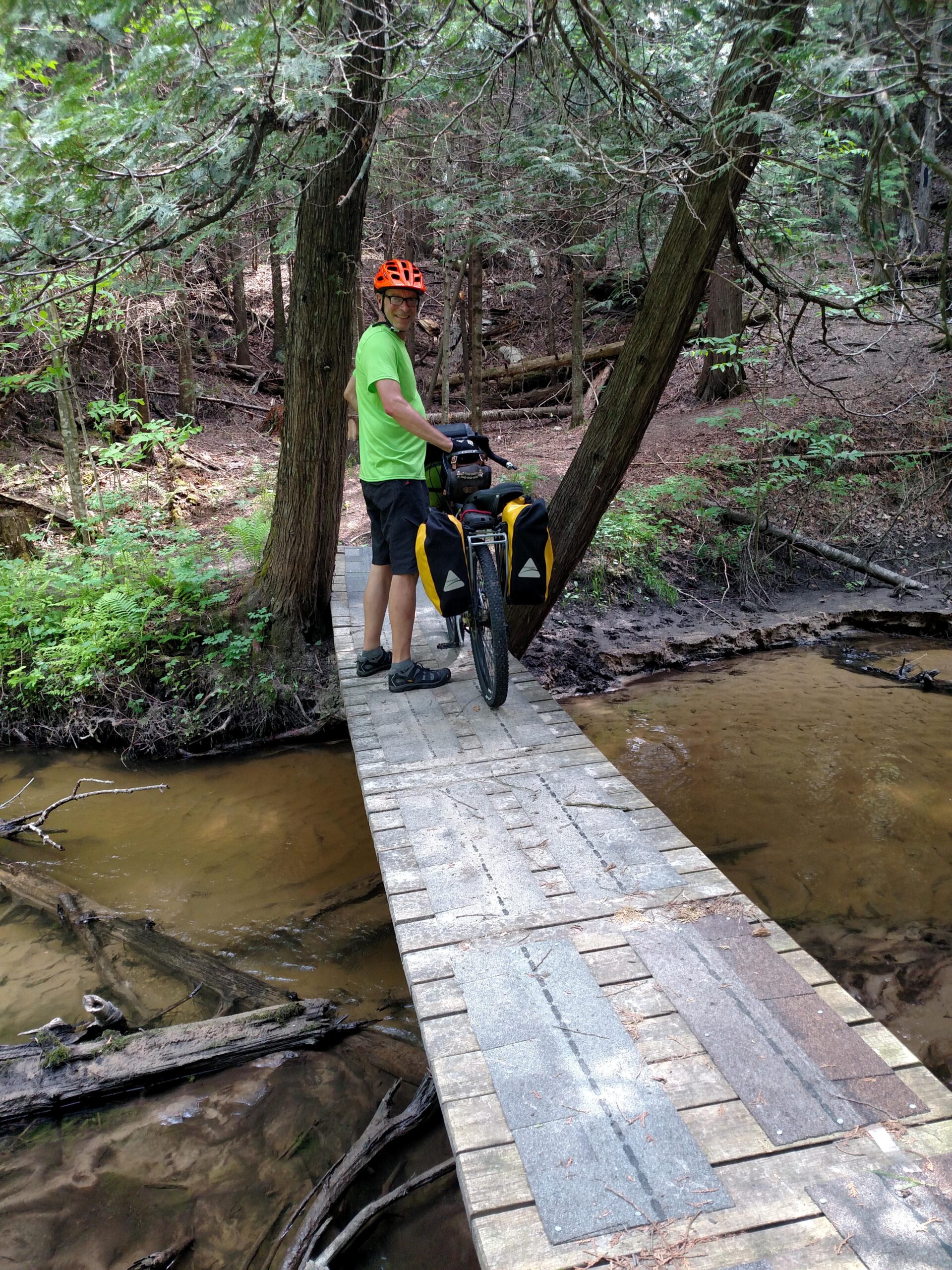 A person wearing an orange helmet and a green shirt stands beside a bicycle with panniers on a narrow wooden bridge in a forested area. The bridge extends over a shallow stream, surrounded by trees and lush greenery. Baxter Bridge east to Fife Lake mountain bike trail.