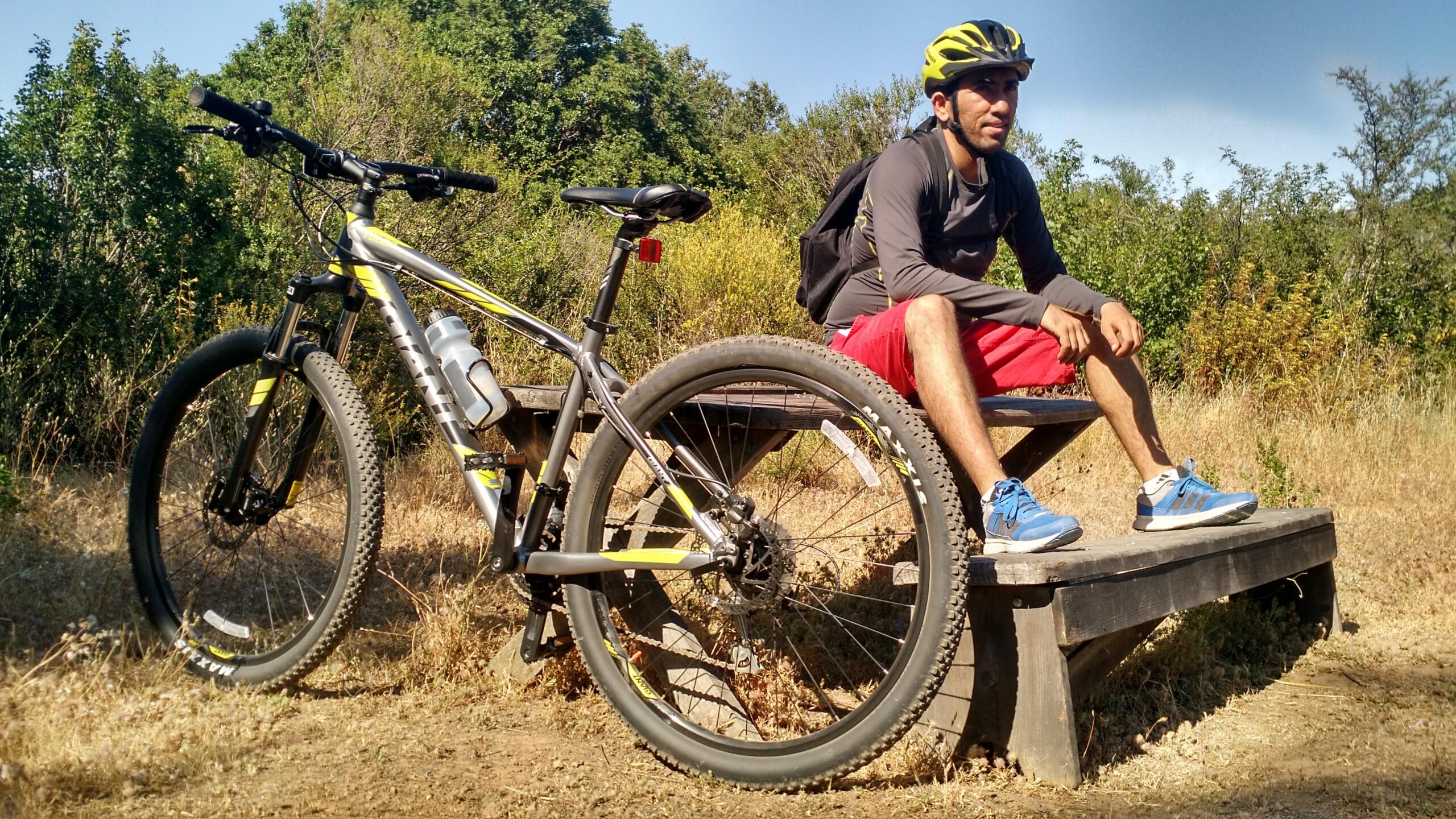 Giant Talon 27.5 4: A cyclist wearing a helmet and a backpack sits on a wooden bench next to a mountain bike. The scene is set in a natural outdoor environment with greenery in the background and dry grass around. The cyclist is dressed in a long-sleeve shirt and red shorts, taking a break while enjoying the surroundings.