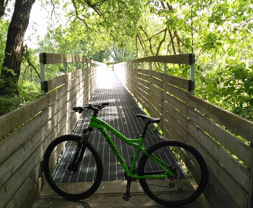 Norco Storm: A green mountain bike is parked on a wooden pathway surrounded by lush green trees and vegetation, leading into a sunny, illuminated area. The path features a slatted design and extends into the distance, inviting exploration.
