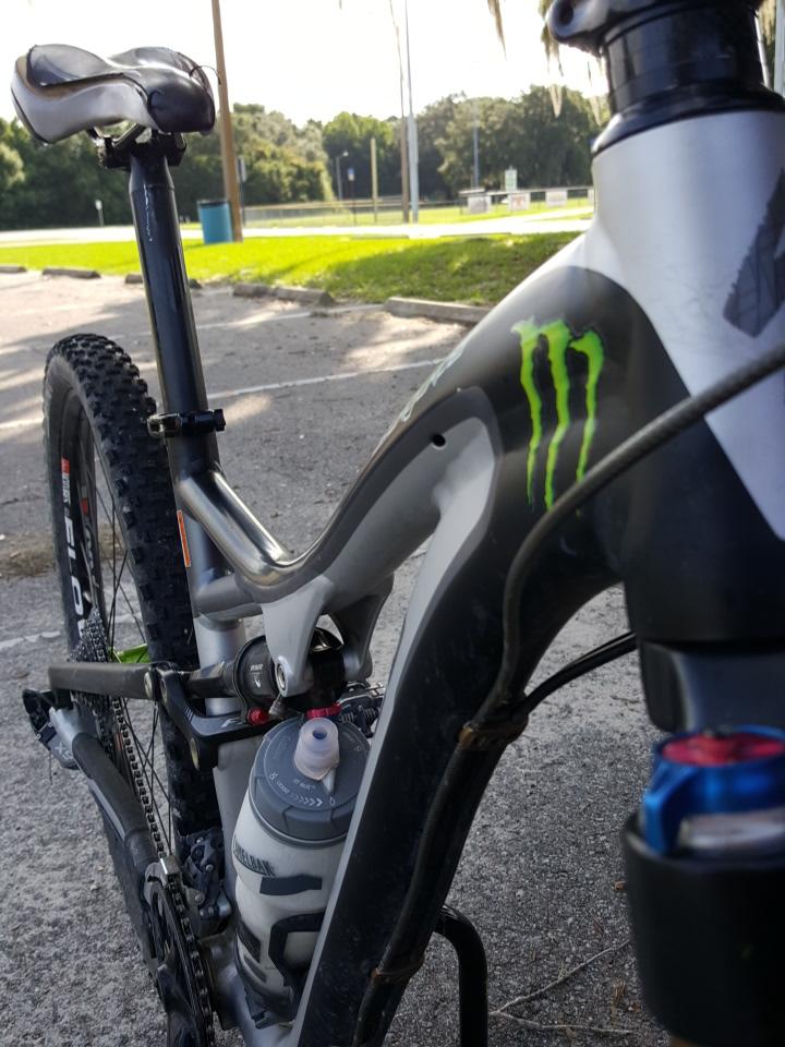 Close-up of a mountain bike showing the frame, seat, and water bottle. The bike features a distinctive graphic with green markings. In the background, a park with grass and a picnic area is visible, indicating an outdoor setting. Loyce E. Harpe Park mountain bike trail.