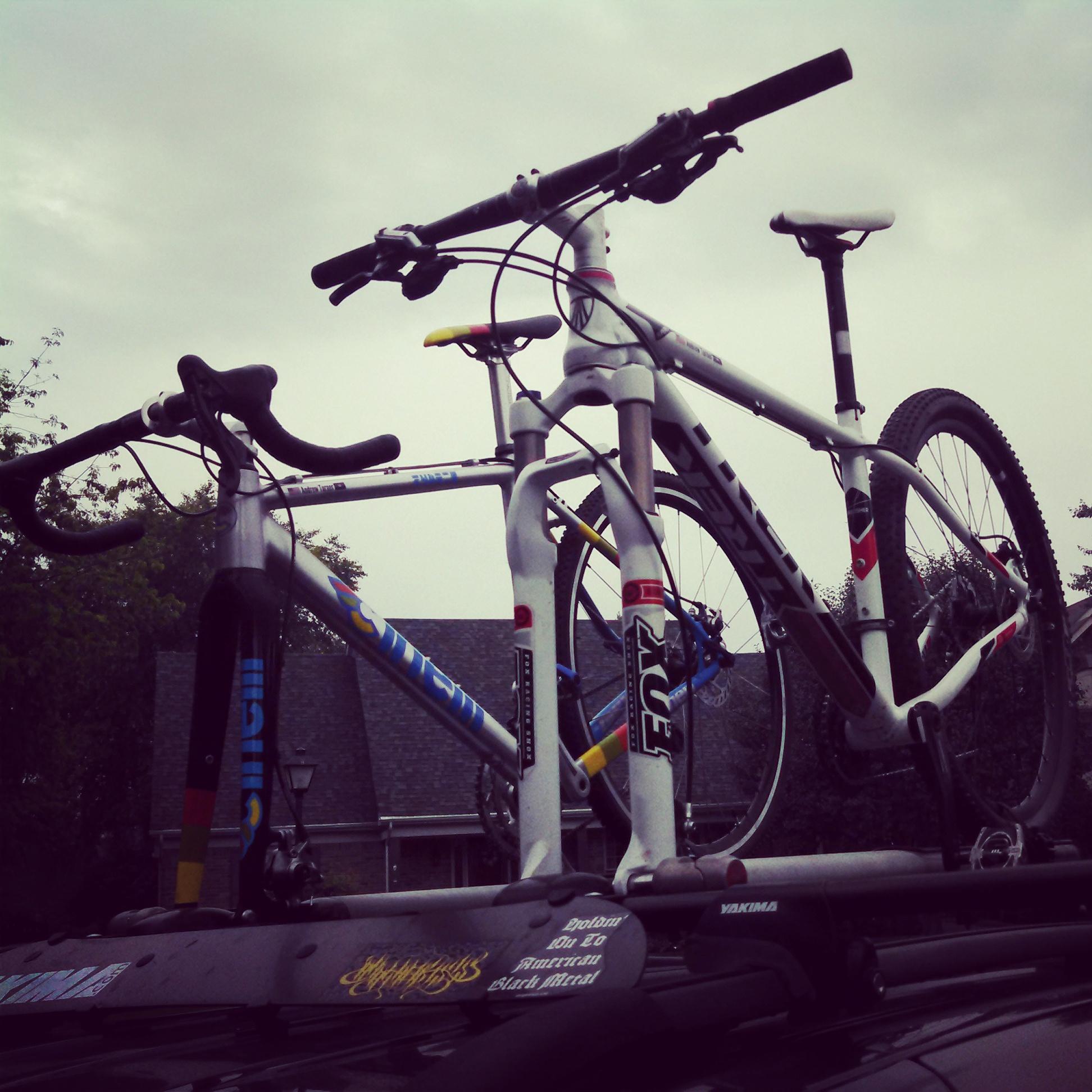 Trek Superfly AL Elite: Two mountain bikes are secured on the roof rack of a car, with a cloudy sky in the background. The bikes are positioned upright, showcasing their handlebars and frames. The rack features additional equipment and branding labels.