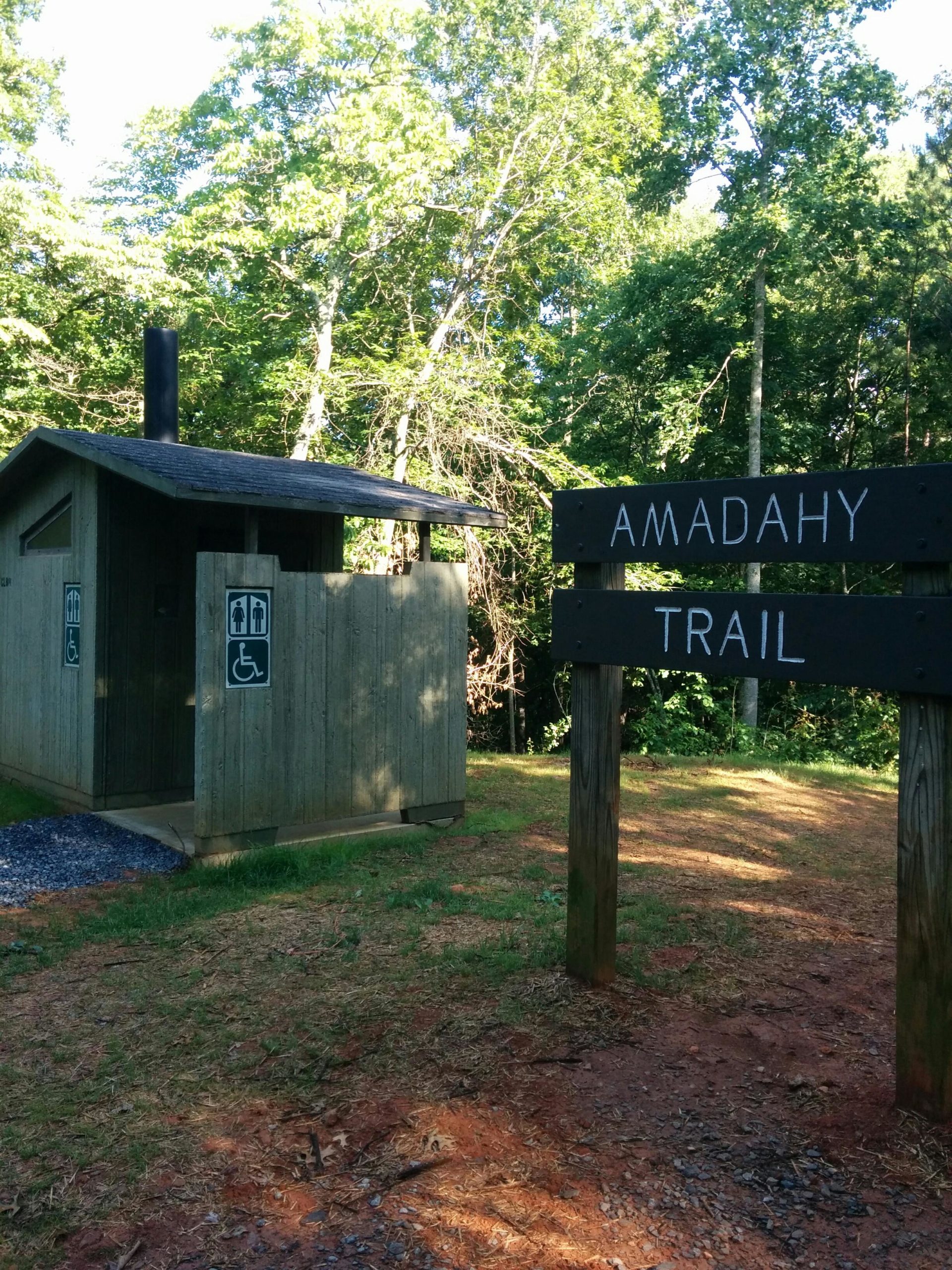 A wooden restroom facility with signs indicating accessible restrooms, situated near the Amadahy Trail. Surrounding the area are lush green trees and a natural landscape, with a dirt path leading to the trail sign. Woodring Branch mountain bike trail.