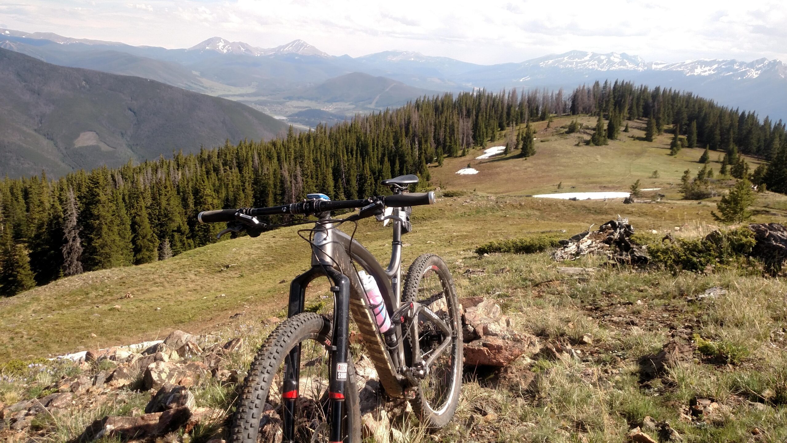 Niner Jet 9 Carbon: A mountain bike resting on rocky terrain, overlooking a scenic view of rolling hills and distant snow-capped mountains under a partly cloudy sky. The foreground features grass and scattered rocks, while dense trees line the background.