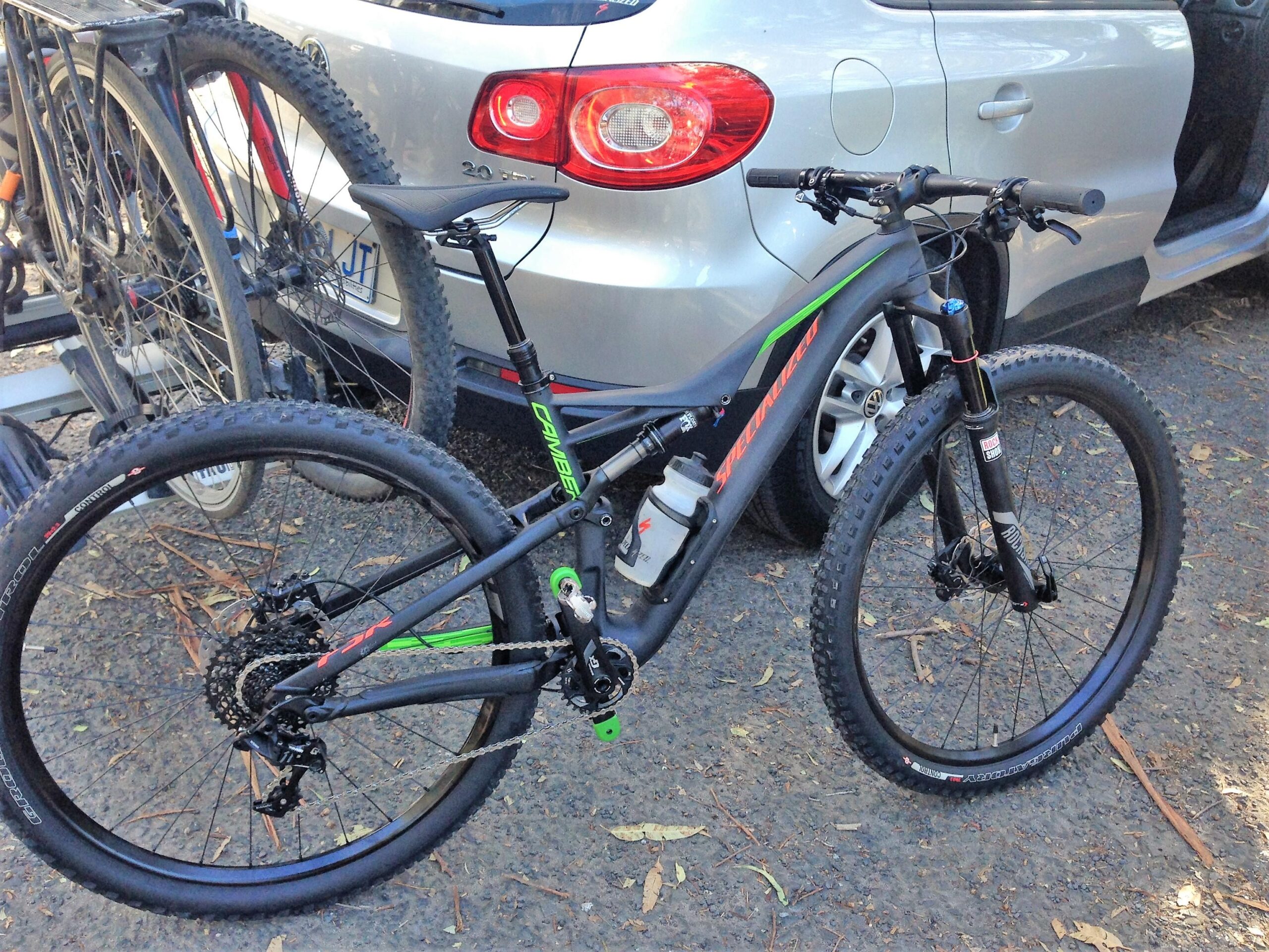 Specialized Camber Comp 29: A modern mountain bike with a black frame accented by green and red details, featuring wide tires and front suspension, parked next to a silver car. The background includes another bicycle rack and a gravel road with scattered leaves.