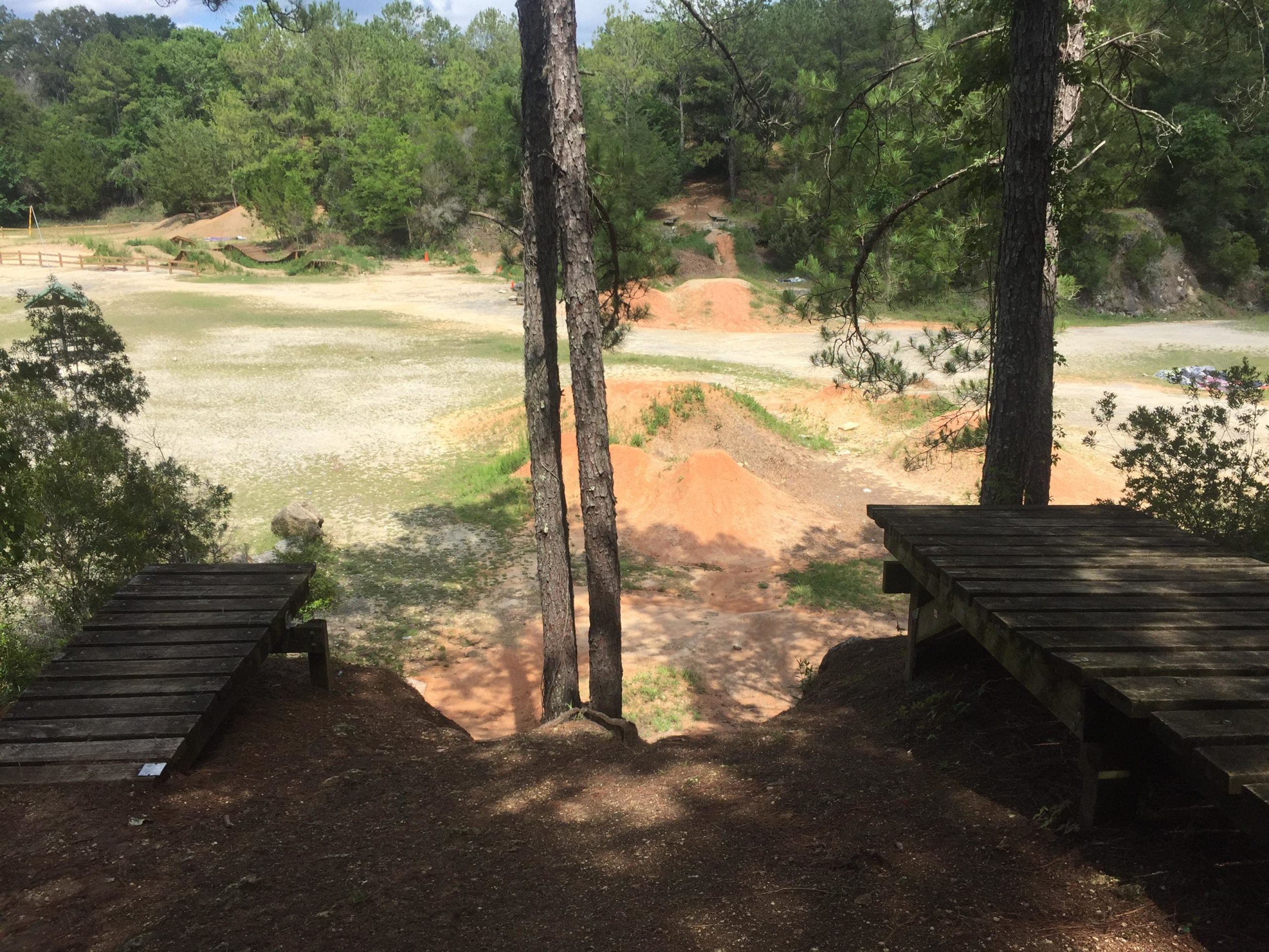 A view of a dirt bike or BMX track with wooden ramps leading down to a sandy area. The landscape features green trees and bushes in the background, with piles of dirt and a flat, open area. The scene is set under a partly cloudy sky, indicating a bright day. Santos mountain bike trail.