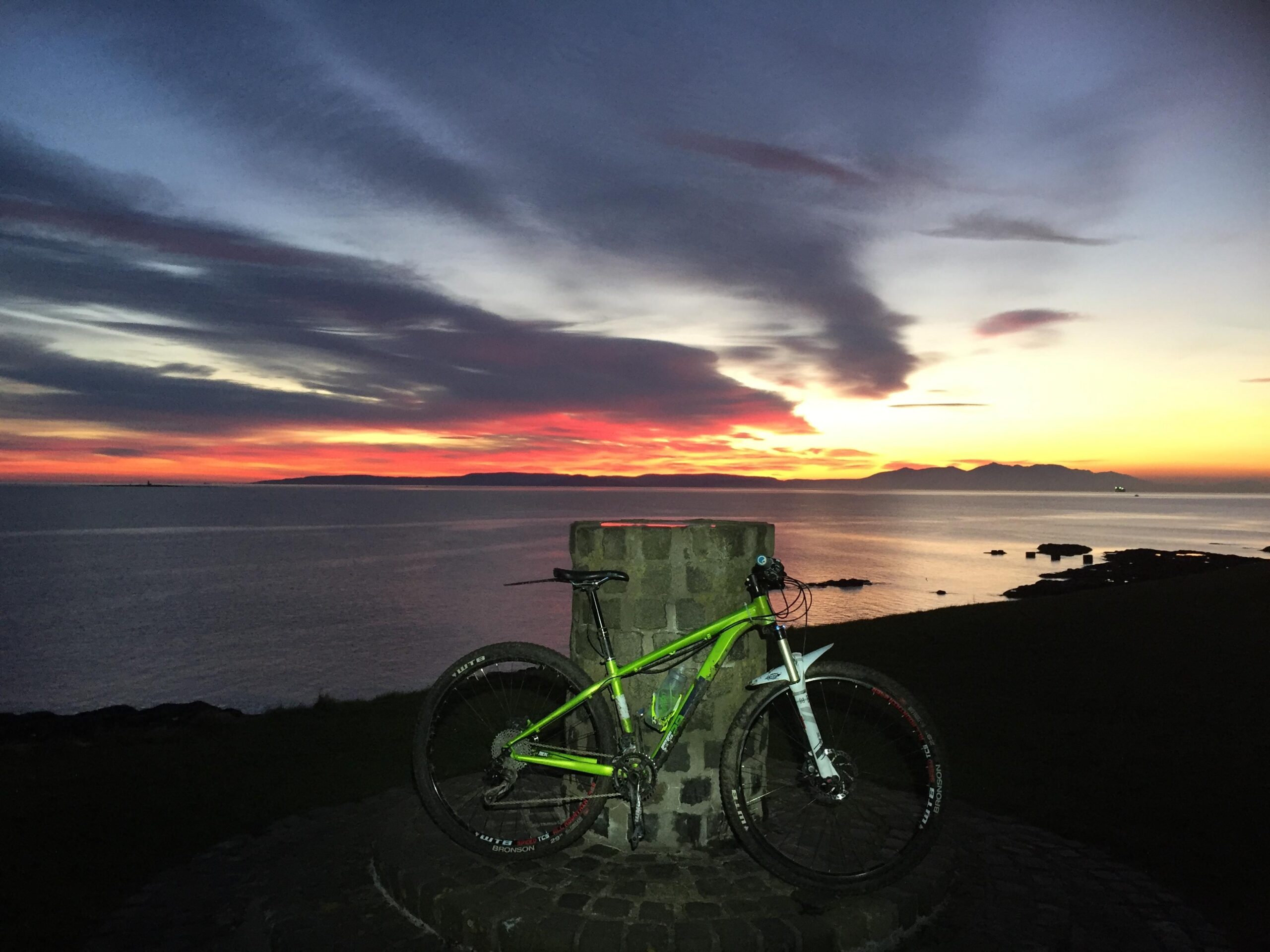 Kinesis FF29: A vibrant sunset over the water, featuring striking orange and purple hues in the sky. In the foreground, a green mountain bike leans against a stone pillar, overlooking the calm sea with distant hills and rocks visible in the background.