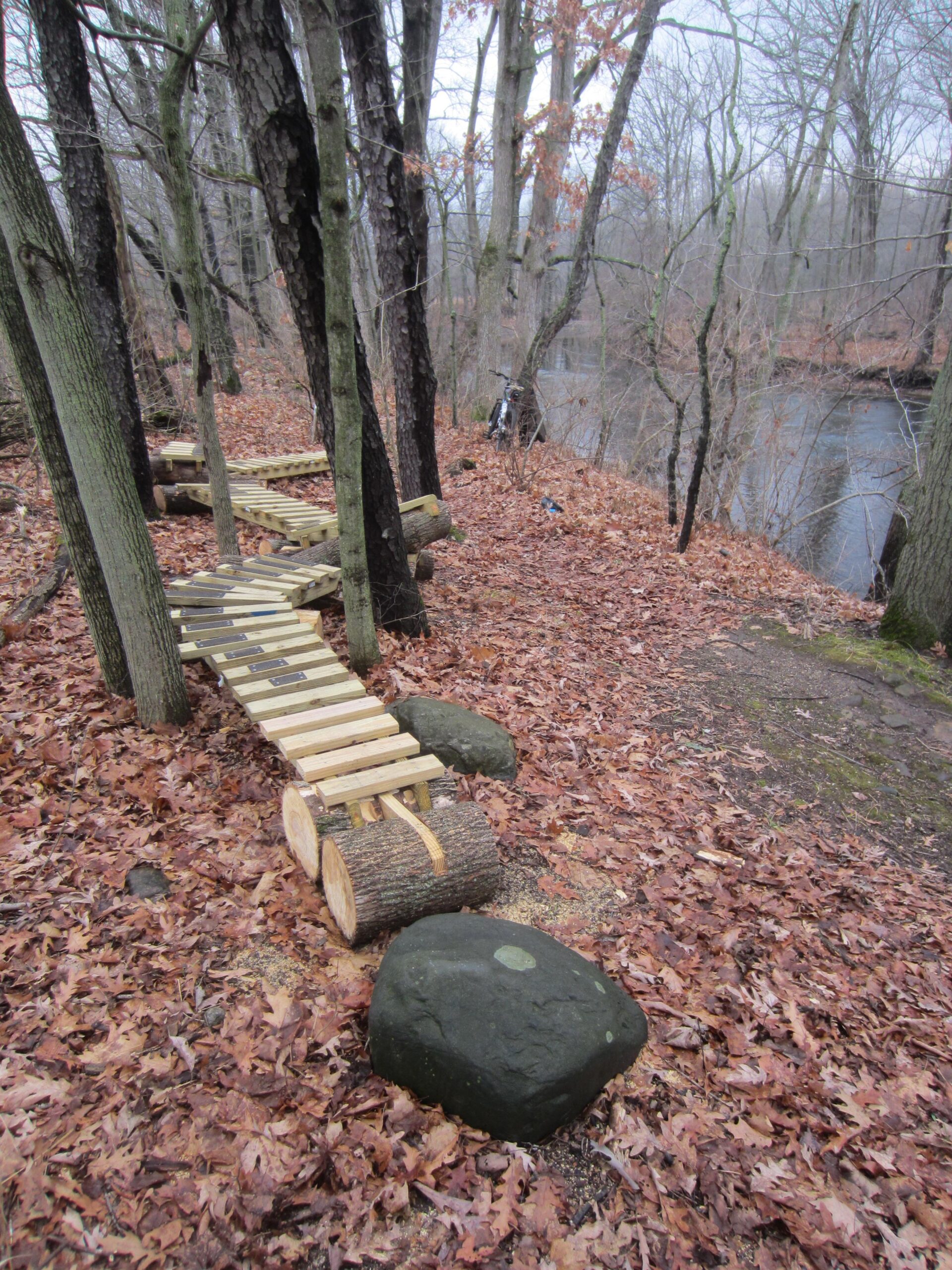 A wooden footbridge made of planks and logs crossing over a pathway covered with fallen leaves, surrounded by trees along the edge of a river. In the background, a bicycle can be seen leaning against a tree. The scene is set in a wooded area during the fall season. Burchfield mountain bike trail.