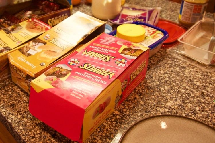 A countertop with various food items, including Honey Stinger product boxes such as Raspberry Protein Chews and Honey Waffles, bags of snacks, and other kitchen items like condiments and a plate.