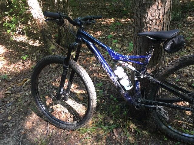 A mountain bike is leaning against a tree in a wooded area with a forest floor covered in leaves. The bike features a blue frame and has a water bottle attached to its frame. Sunlight filters through the trees, creating a dappled light effect on the scene. Rosaryville State Park mountain bike trail.