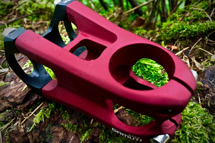 A close-up image of a red bicycle stem resting on a bed of moss and bark. The stem features a modern design with a glossy finish and is partially wet, likely from recent rain. The background includes soft green moss and blurred foliage, creating a natural outdoor setting.